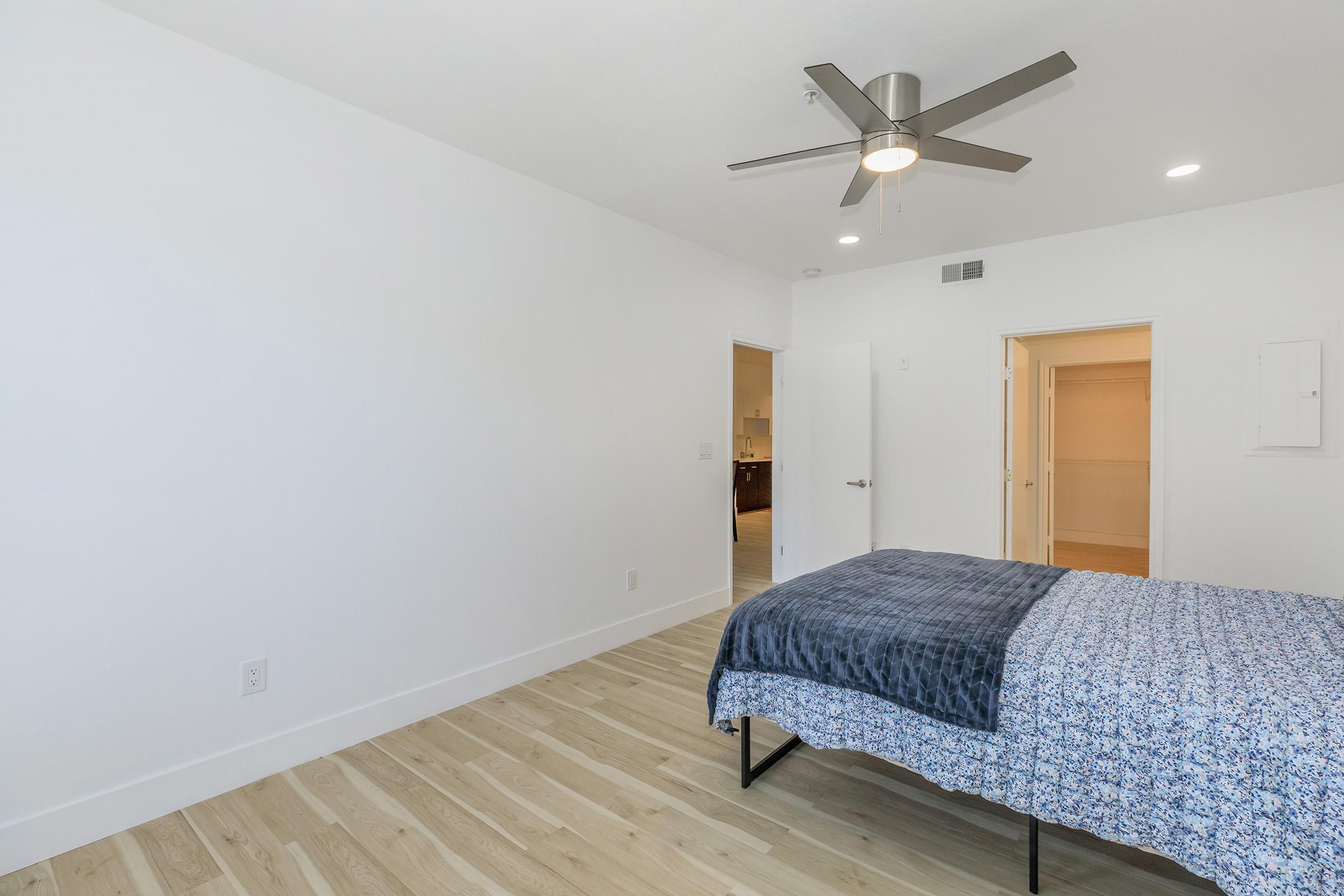 A bright, spacious bedroom featuring a ceiling fan, light-colored walls, and light wood flooring. A neatly made bed with a blue patterned comforter is positioned in the center. The room includes a white door leading to an adjacent space, giving a modern and minimalist vibe.