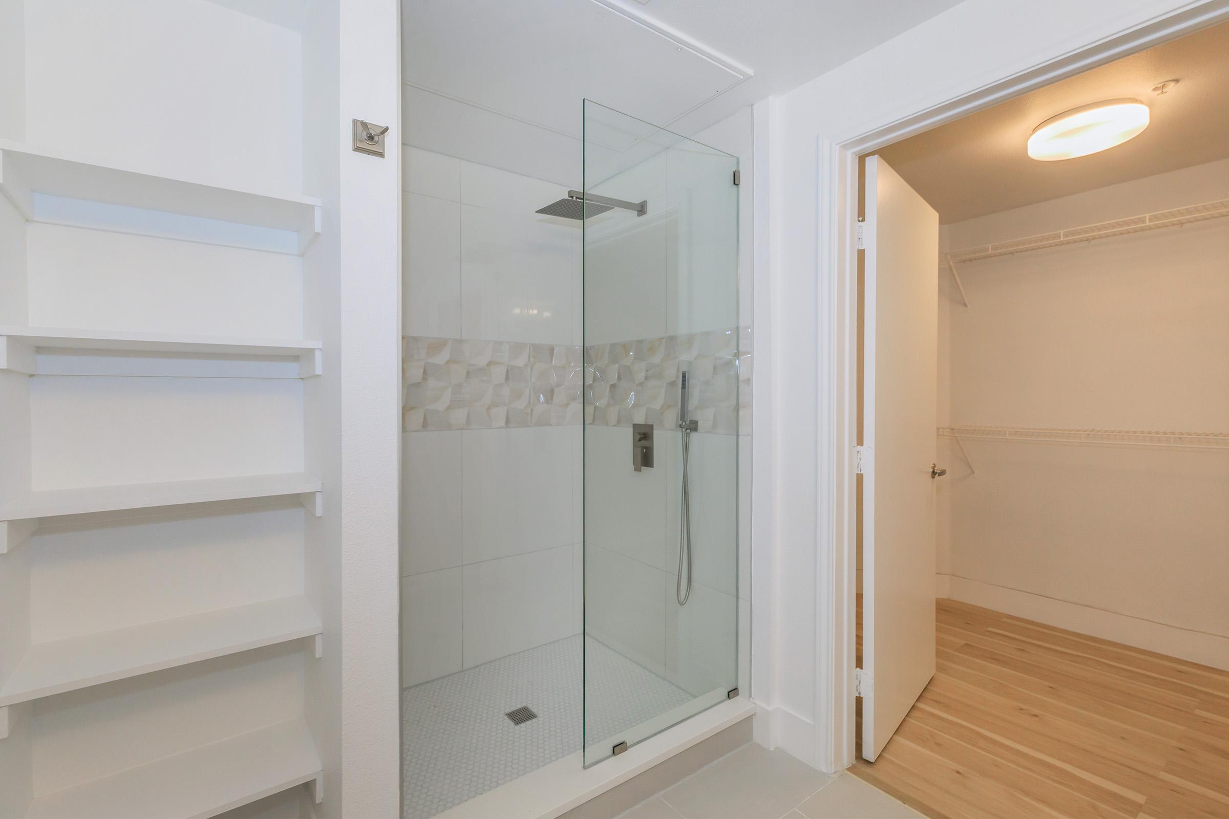 A modern bathroom featuring a glass-enclosed shower with a textured tile accent and a handheld showerhead. Adjacent to the shower is an open closet space with built-in shelves and a doorway leading to a well-lit area with wooden flooring. The walls are painted in a bright, neutral color.