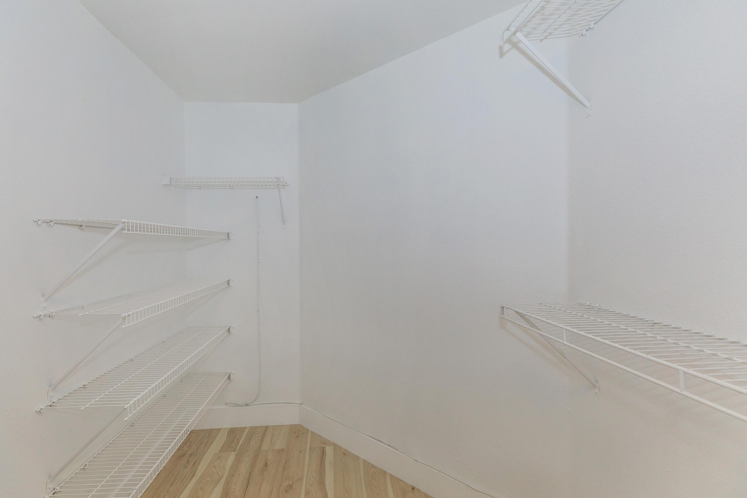 An empty closet featuring white wire shelves arranged along the walls. The floor is light-colored wood. The room has plain white walls and a stark, minimalistic design, emphasizing the spaciousness of the area.
