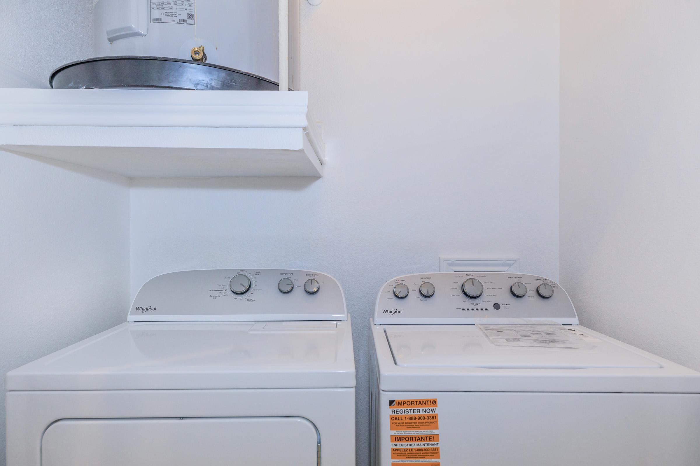 A small laundry area featuring a white washing machine and a matching dryer side by side. Above the appliances is a shelf with a circular storage container. The walls are painted white, creating a clean and bright appearance.