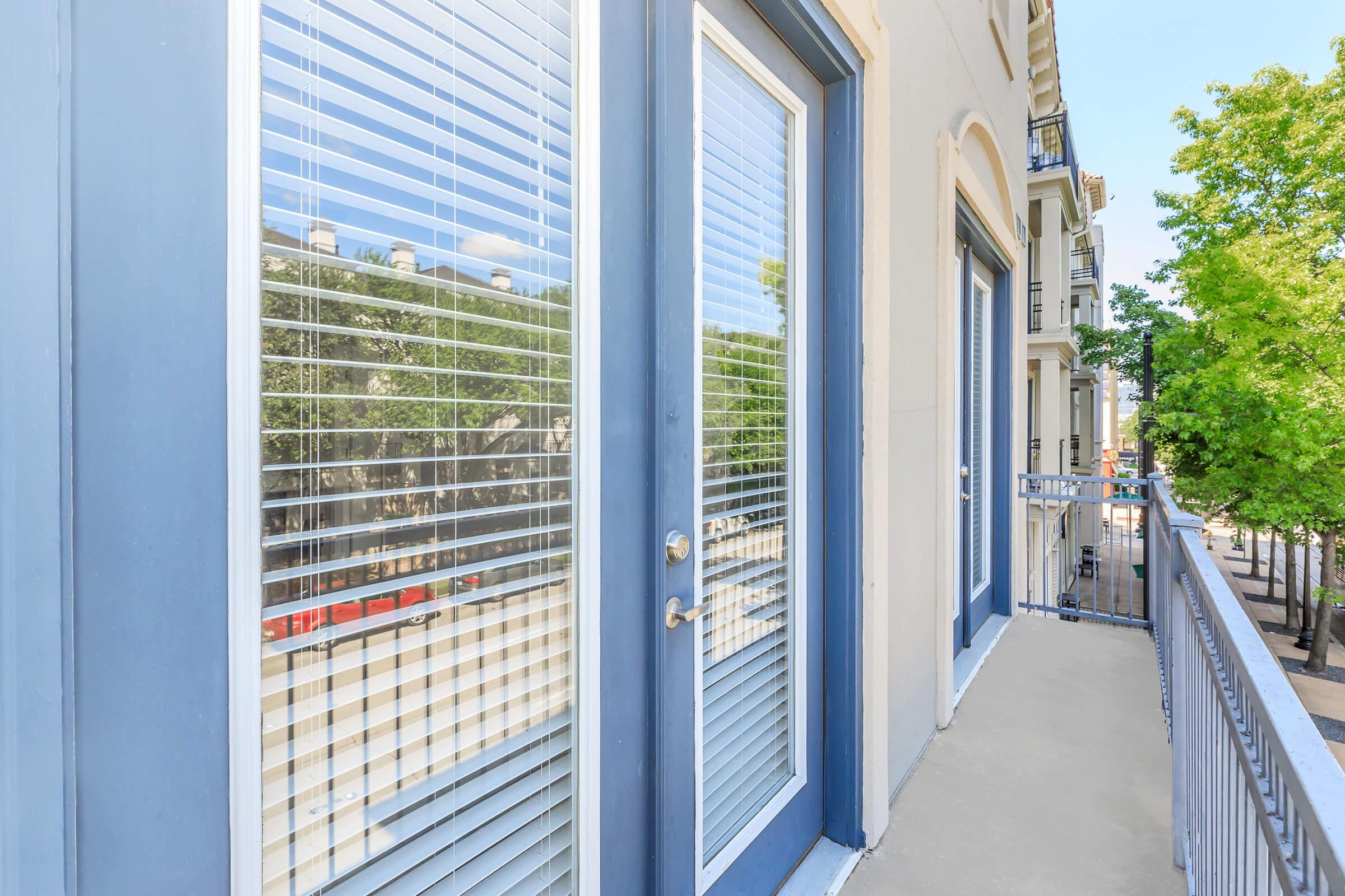 A balcony view featuring two large glass doors with blinds. The doors frame a reflection of trees and buildings outside. A railing runs along the edge of the balcony, and the flooring is smooth concrete, suggesting a modern apartment setting. The scene is bright with natural light.