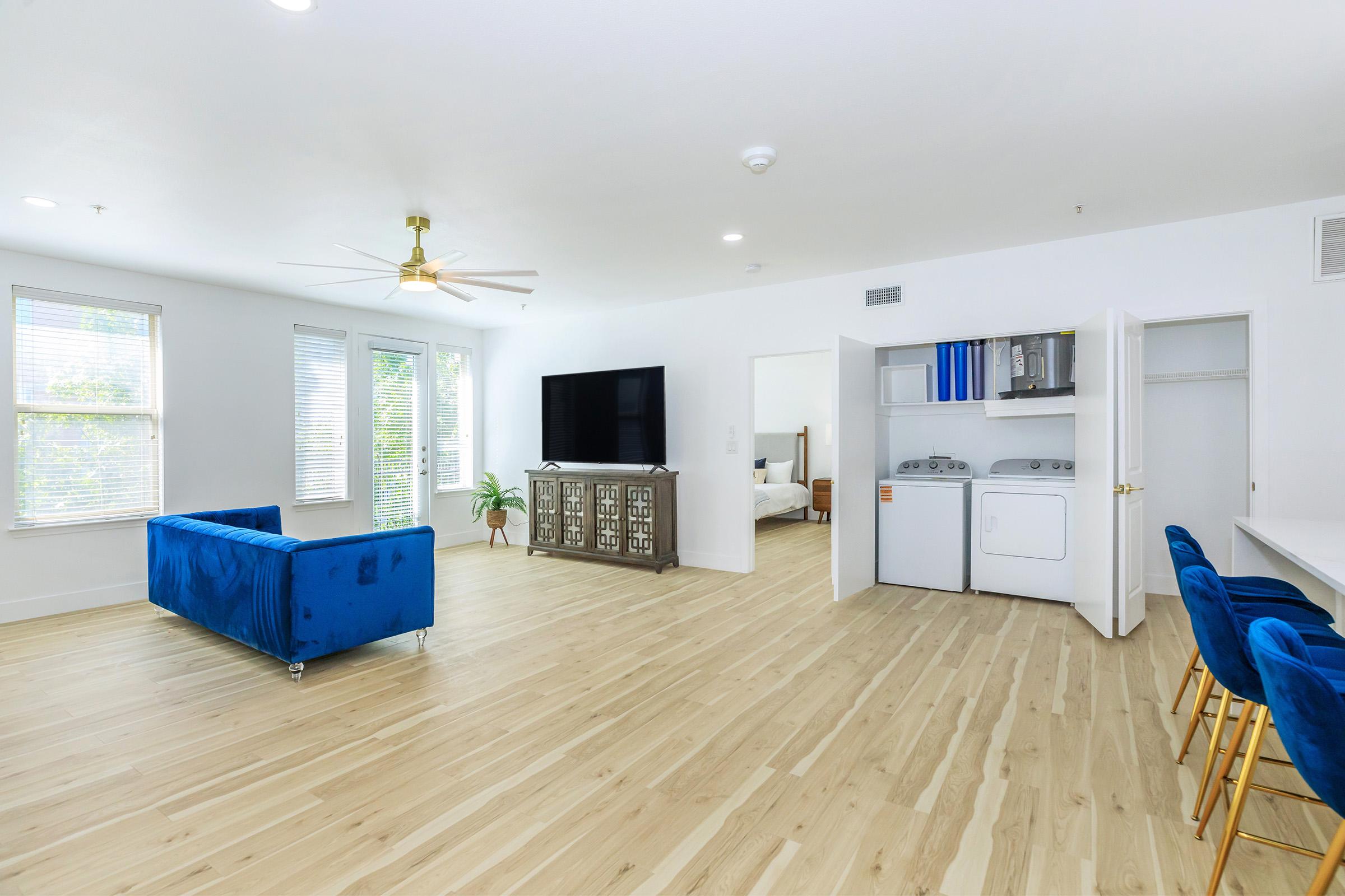 Bright and modern living space featuring a blue sofa, a large TV, and a stylish cabinet. In the background, there is a laundry area with stacked washer and dryer units. Natural light shines through the windows, enhancing the light wood flooring and creating a welcoming atmosphere.