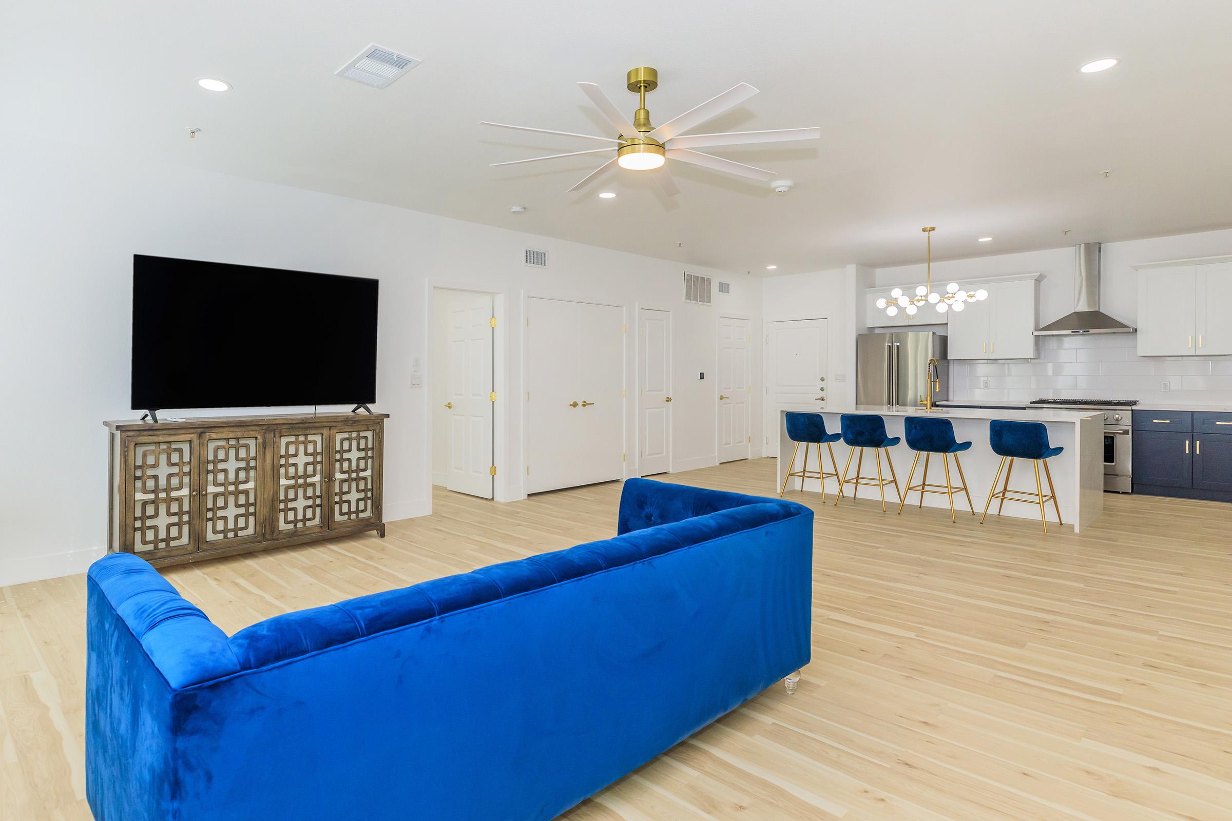 A modern living space featuring a blue velvet sofa, a large flat-screen TV, and a stylish cabinet. The open kitchen with bar seating has white cabinetry and stainless steel appliances. Light hardwood flooring and a ceiling fan add to the contemporary decor, with bright white walls enhancing the spacious feel.