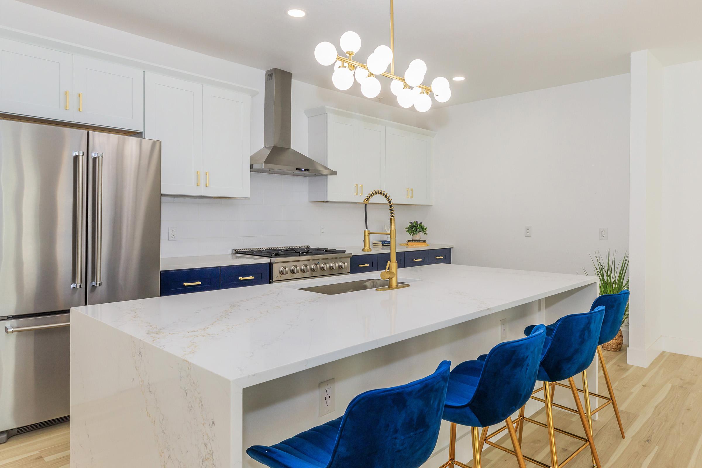 A modern kitchen featuring a large white marble island with a built-in sink, surrounded by blue velvet barstools. The kitchen has white cabinets, a stainless steel refrigerator, and a gas stove with a metal range hood. A stylish chandelier hangs from the ceiling, providing warm lighting.