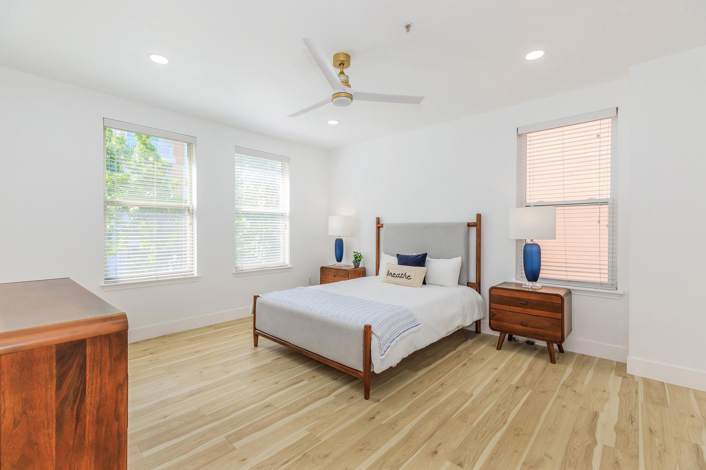 A bright, modern bedroom featuring a cozy bed with a light gray blanket, two bedside tables with blue lamps, and a wooden dresser. The room has large windows letting in natural light, white walls, and a ceiling fan, creating a spacious and inviting atmosphere.