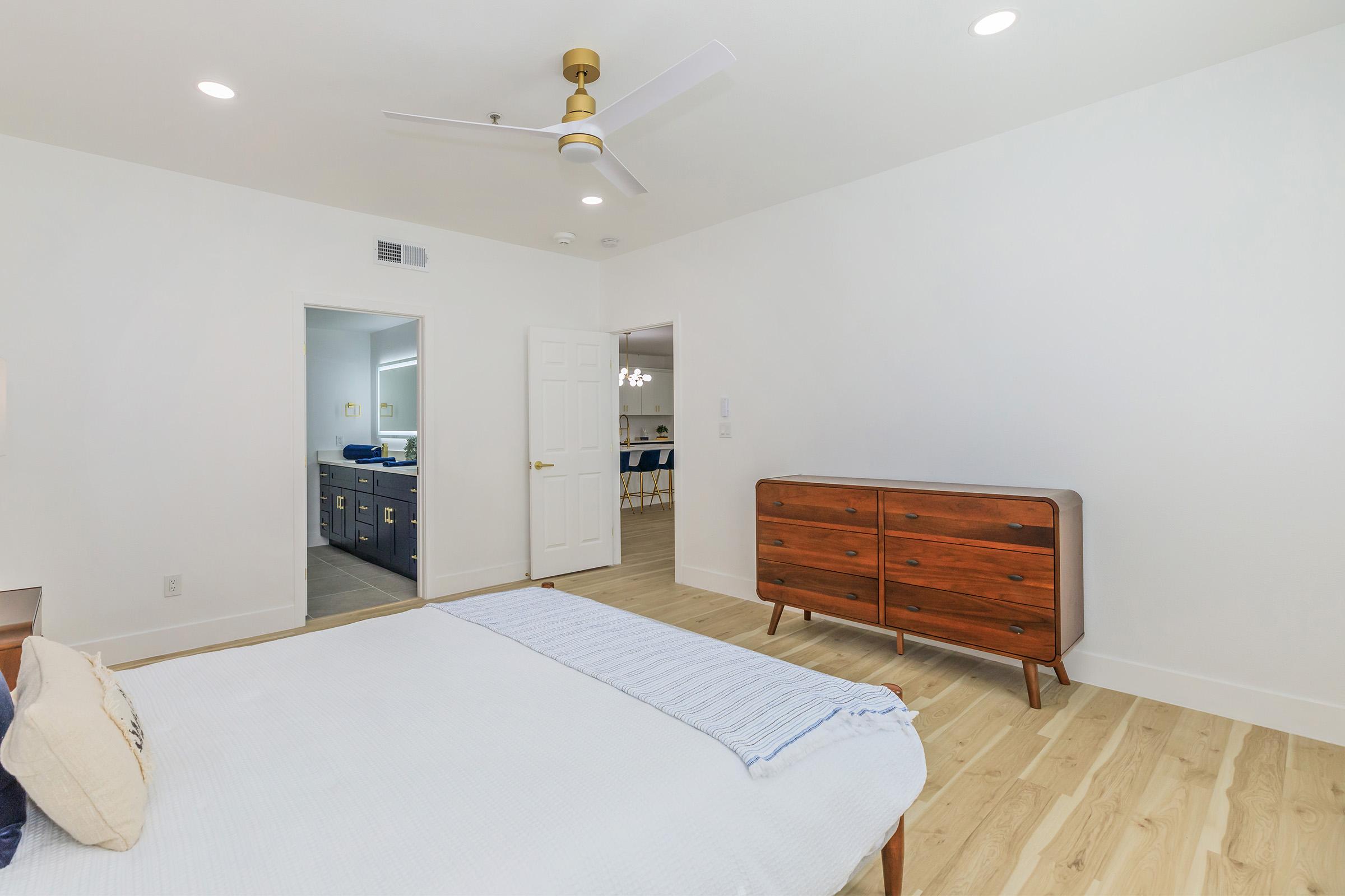 A minimalist bedroom featuring a white wall, wooden dresser, and a bed with a light-colored blanket. A ceiling fan is visible, and an open door leads to another room with blue cabinets. The flooring is light wood, creating a bright and airy atmosphere.