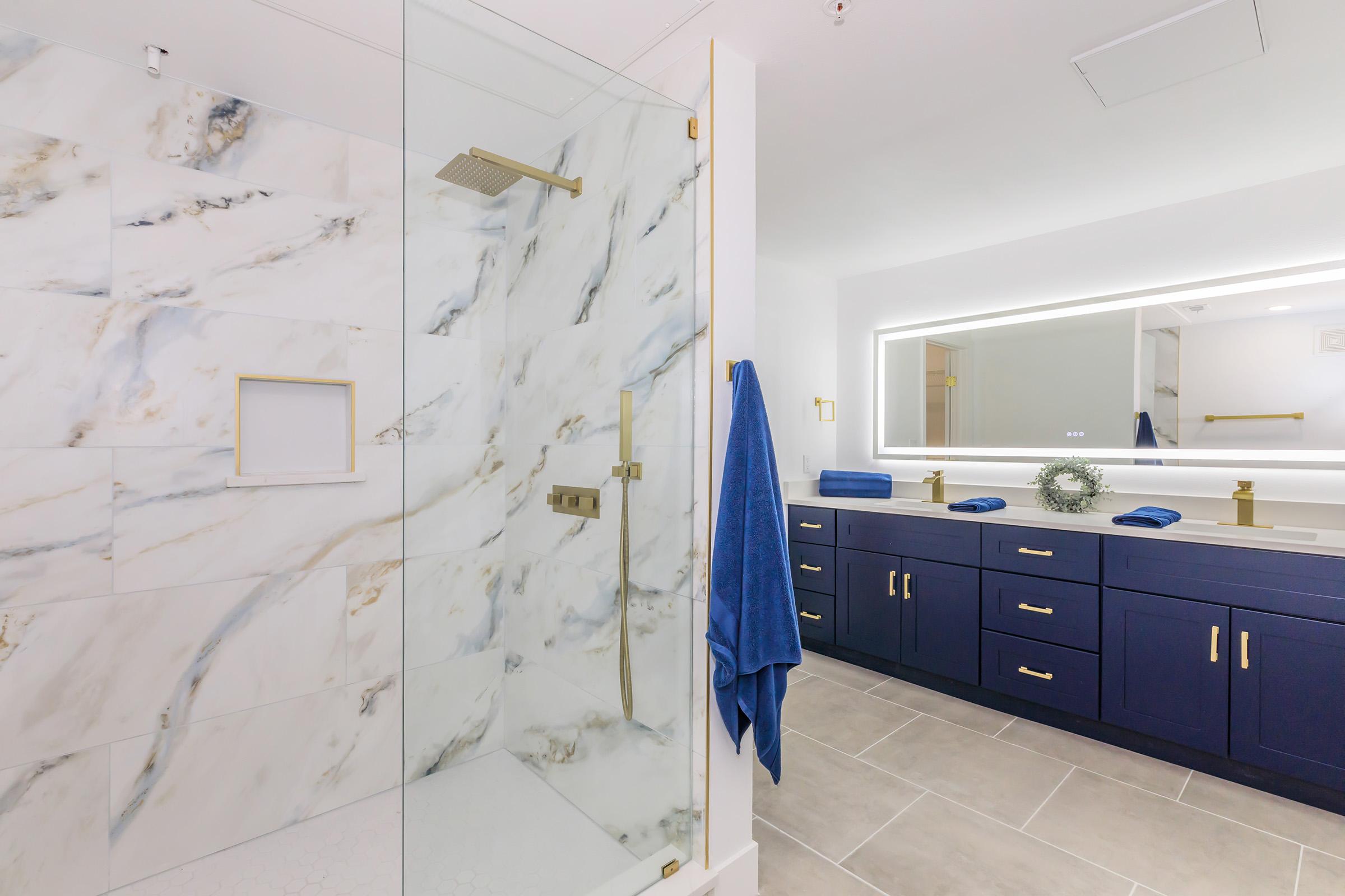 A modern bathroom featuring a glass shower with marble walls, a sleek vanity with dark cabinetry, a large illuminated mirror, and decorative blue towels. The space has a bright and contemporary aesthetic with elegant fixtures.