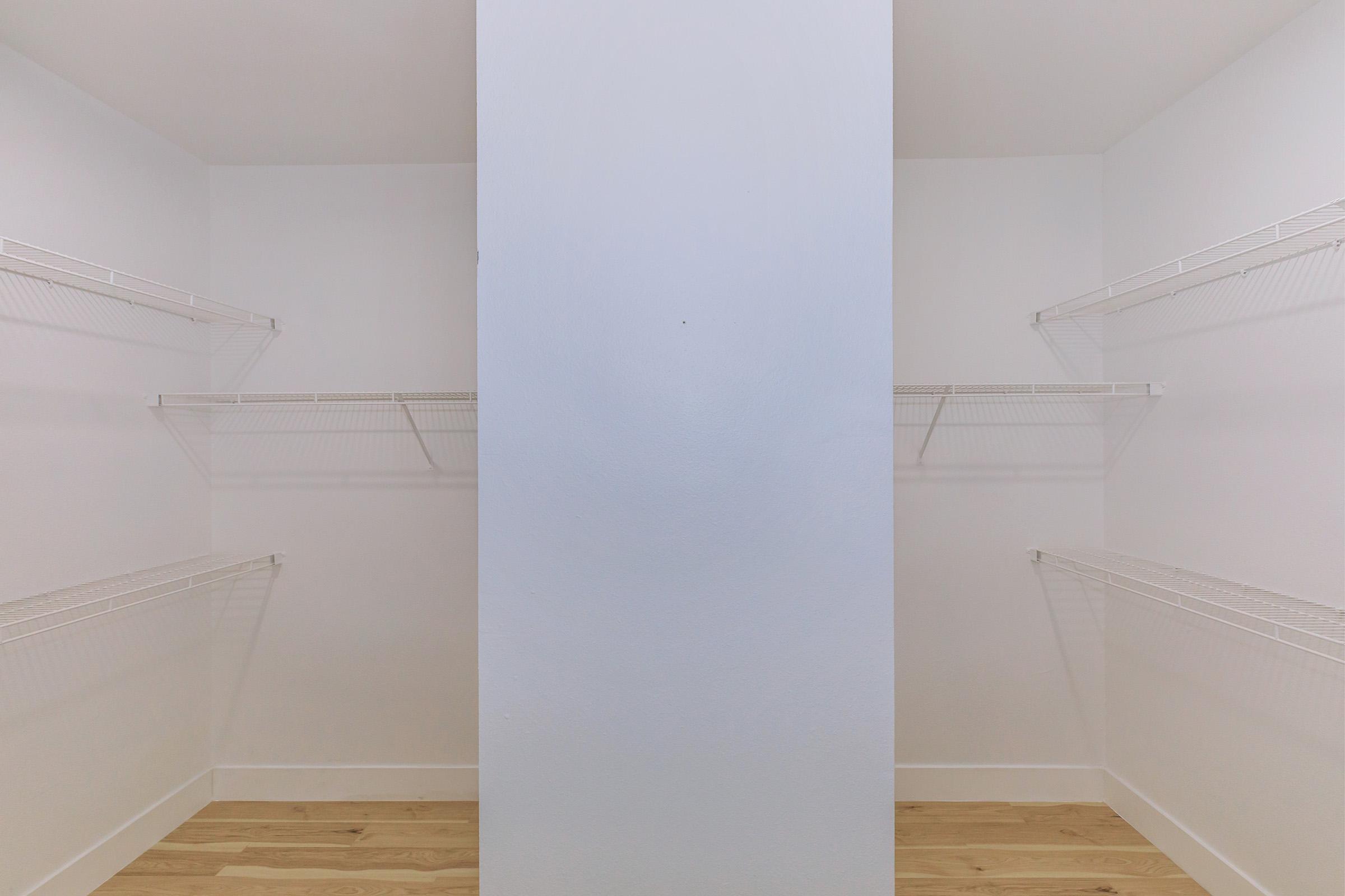 A spacious walk-in closet featuring white walls and a central partition. The closet has several wire shelves installed along the sides, providing ample storage space. The floor is made of light-colored wood, enhancing the clean and minimalist look of the area.