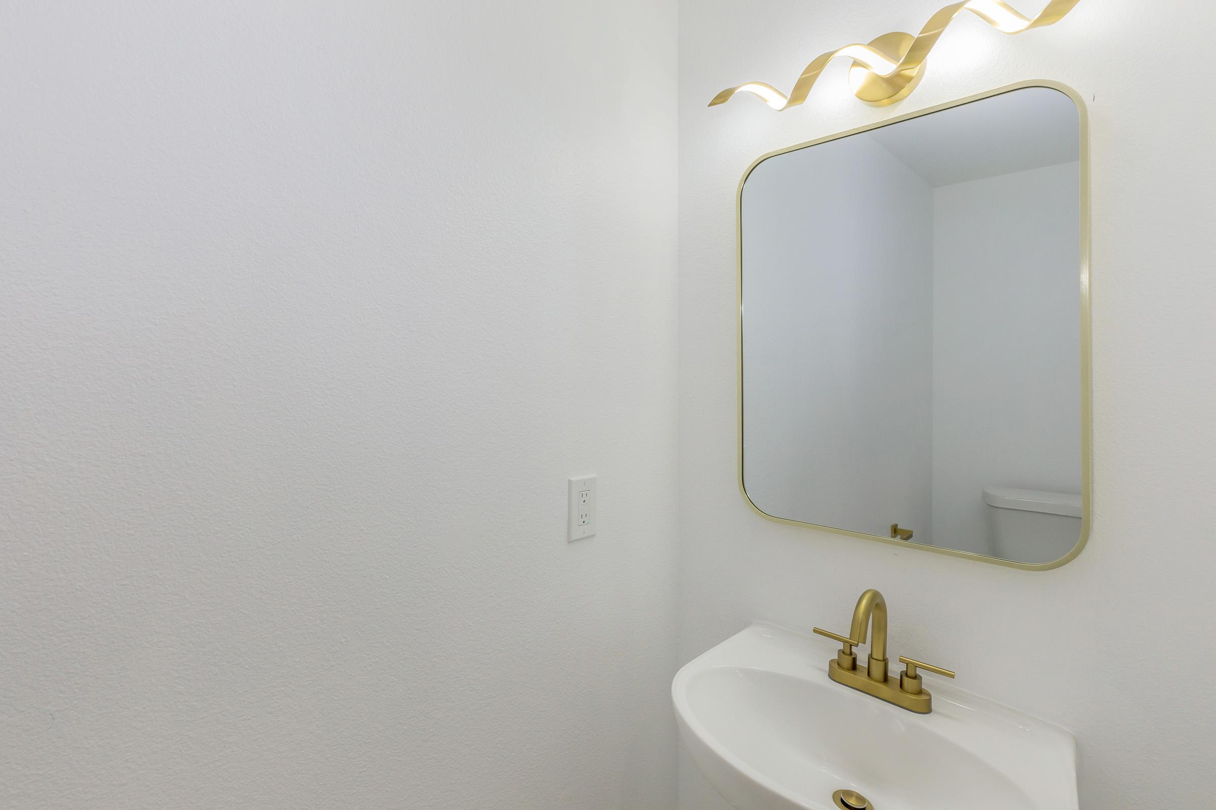 A modern bathroom featuring a white wall, a sleek white sink with a gold faucet, and a large mirror with a decorative gold frame. The space has minimalist decor, and a light fixture above the mirror casts a soft glow, enhancing the clean aesthetic. A glimpse of a toilet is visible in the background.