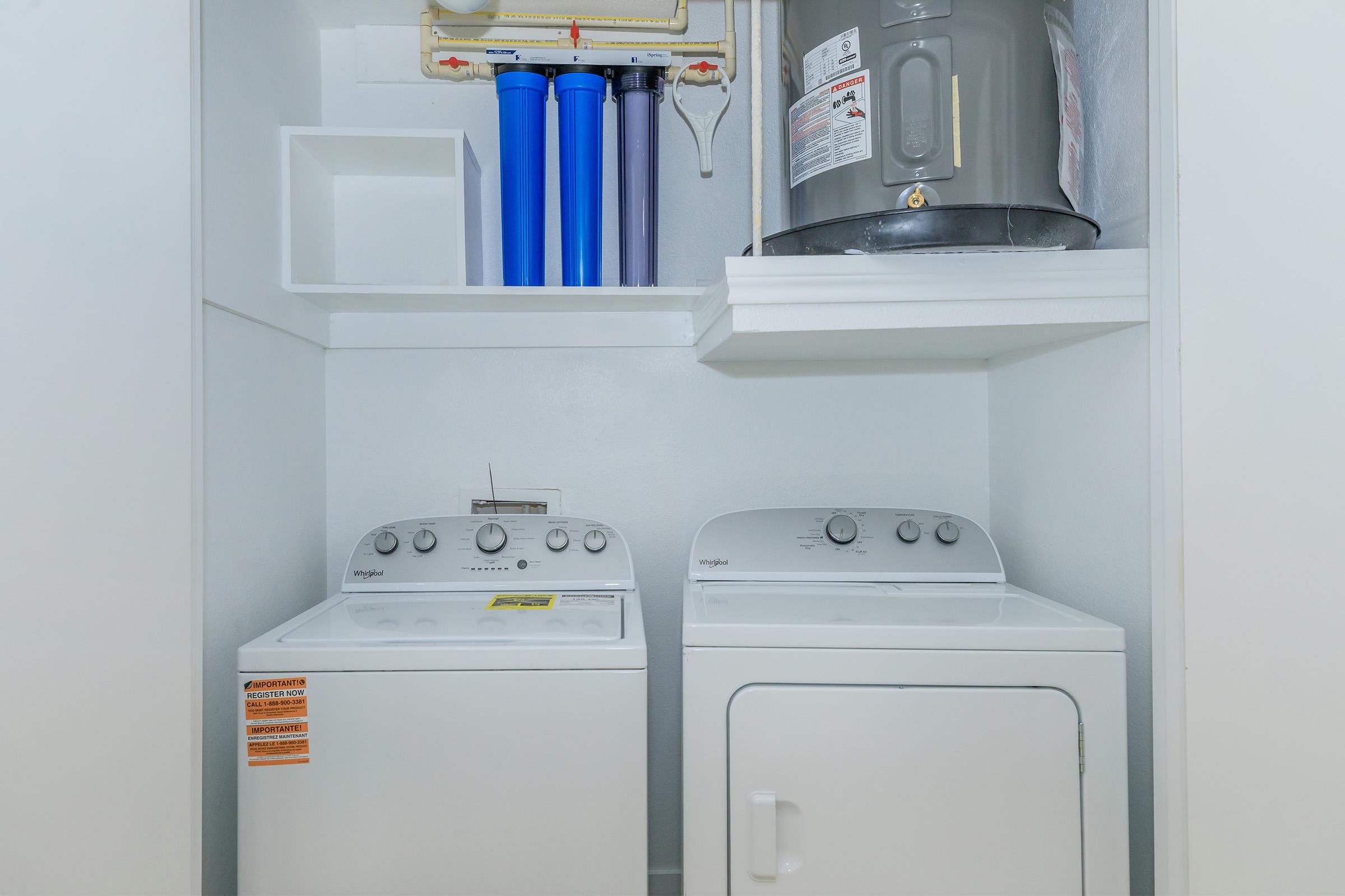 A clean laundry area featuring a white washer and dryer, stacked against a wall. Above them is a water heater and several blue water filtration containers, with a shelf for additional storage. The space has a bright, organized appearance.