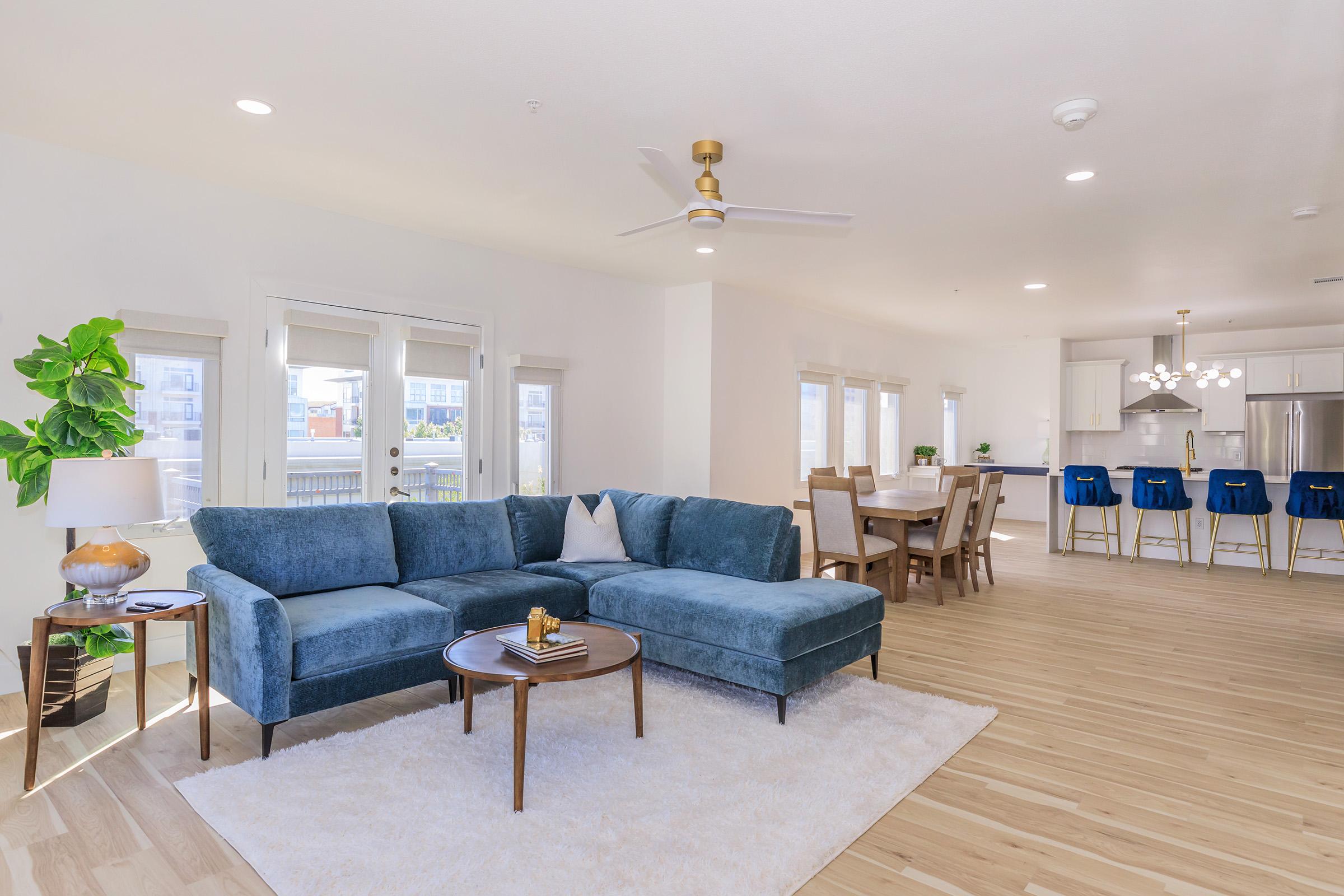 A spacious modern living area featuring a blue sectional sofa, a round coffee table, and a white area rug. Large windows allow natural light, and a dining area with a wooden table and blue bar stools is visible in the background. A plant adds a touch of greenery to the welcoming, open layout.