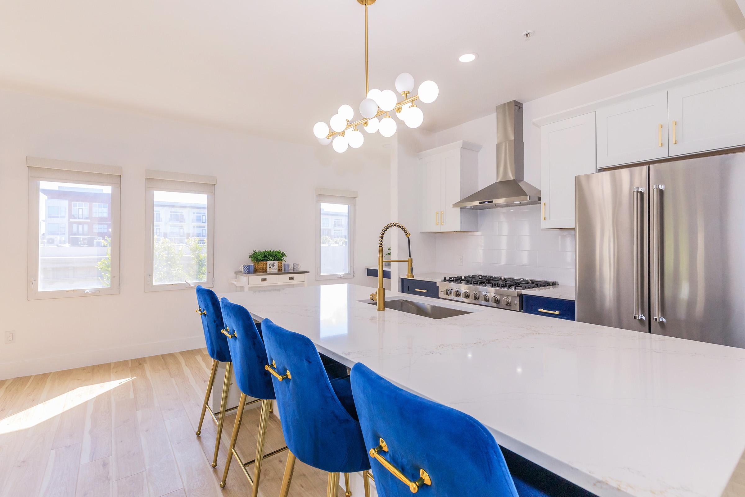 A modern kitchen featuring a large white marble island with gold accents and four blue velvet stools. The kitchen has white cabinets, stainless steel appliances, and a stylish chandelier. Natural light flows in through three windows, enhancing the contemporary atmosphere.