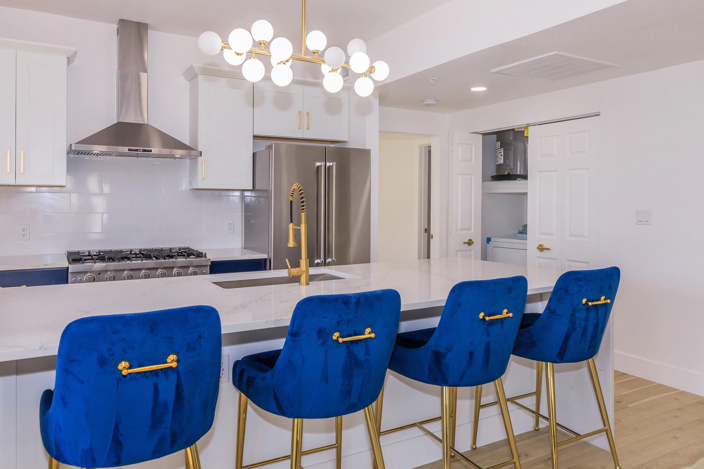 A modern kitchen featuring a large white marble island with four blue velvet bar stools, a stylish gold faucet, and a statement chandelier. Stainless steel appliances, including a refrigerator and a stove, are visible, along with white cabinets and light-colored flooring.
