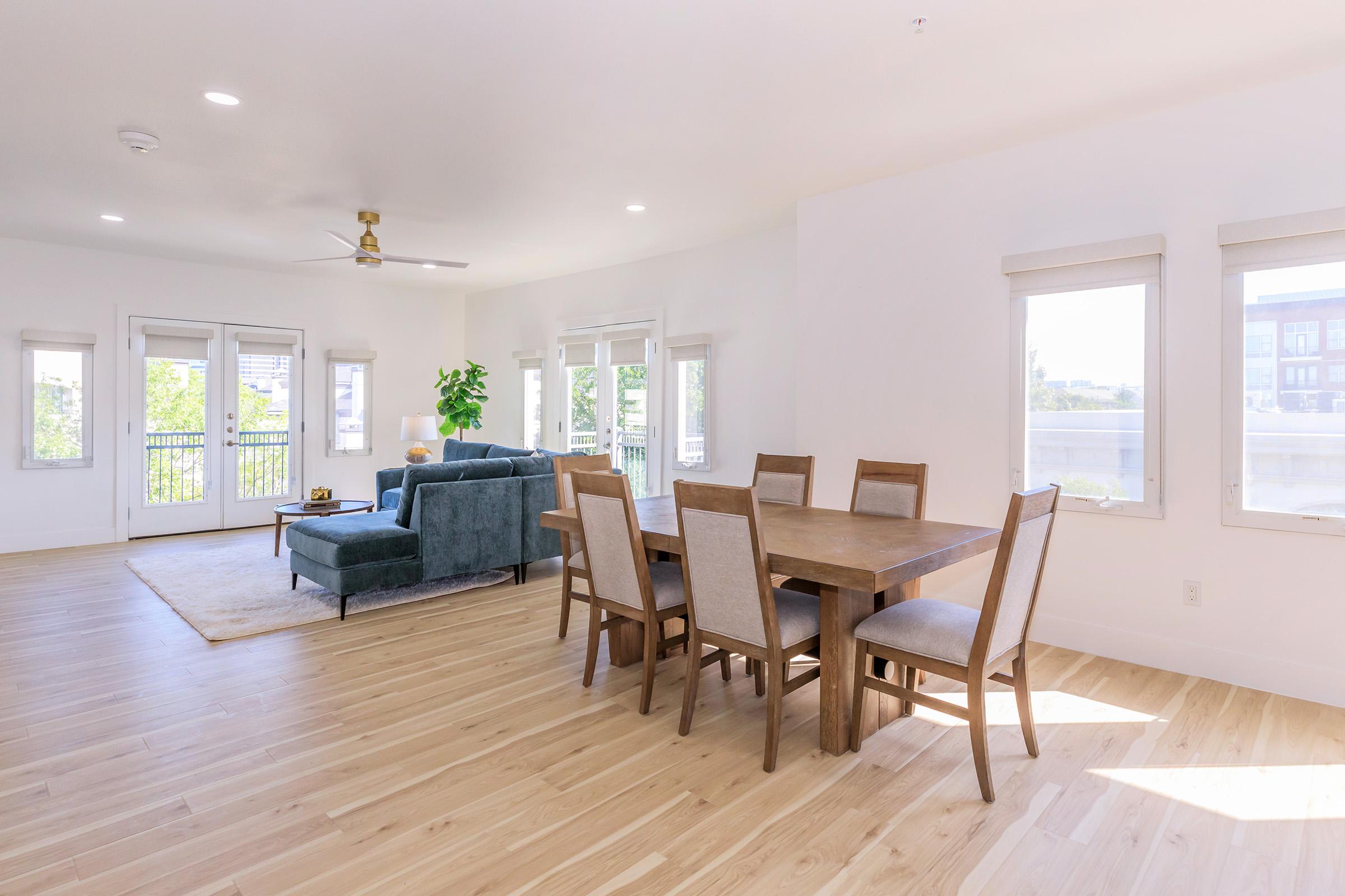 Bright, open living space featuring a cozy blue sofa, a wooden dining table with six chairs, and large windows allowing natural light. The light-colored wooden floor adds warmth, while a green plant adds a touch of freshness to the modern, minimalist design.