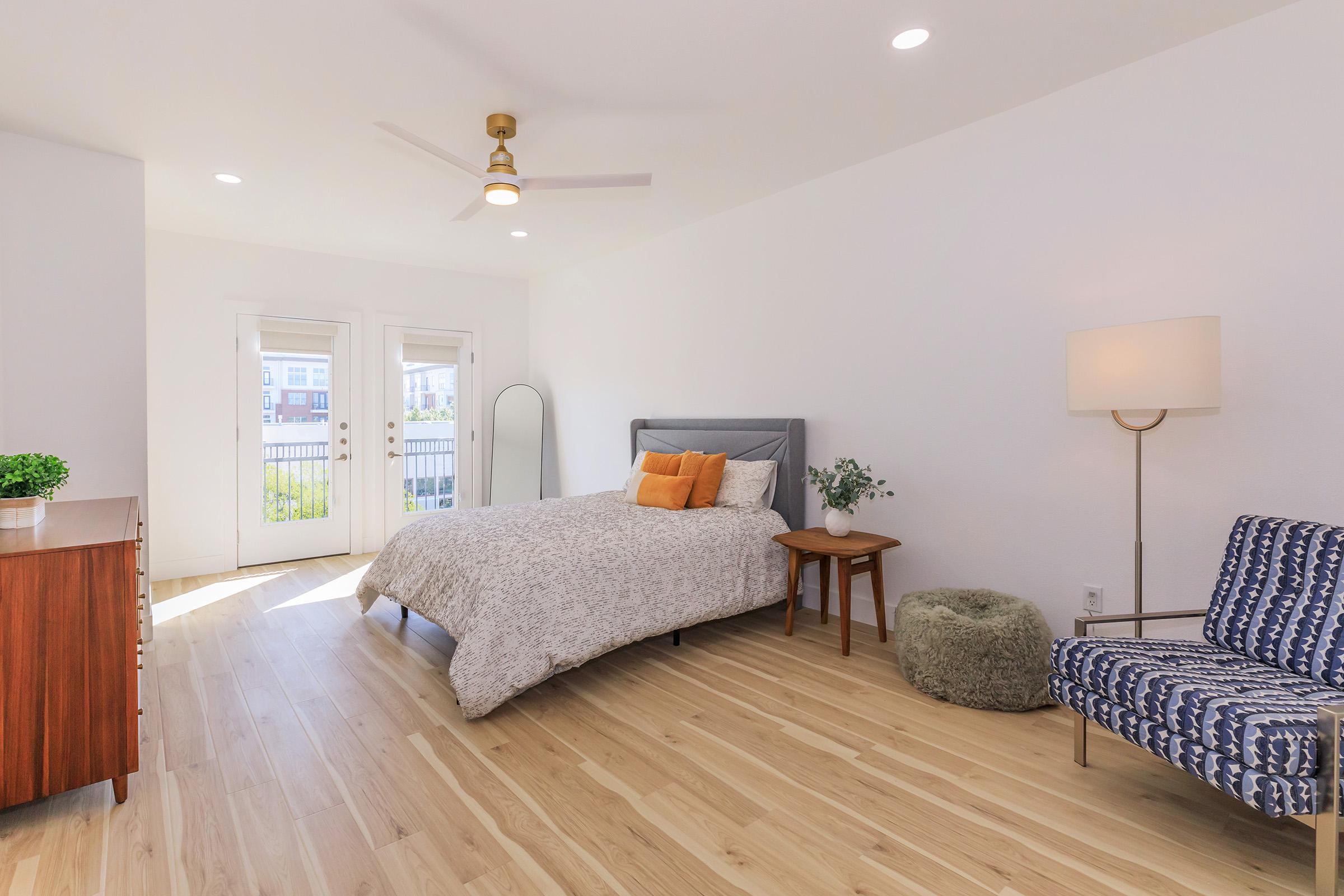 A bright, modern bedroom featuring a queen-sized bed with decorative pillows, a wooden nightstand, and a cozy armchair. Large sliding glass doors open to a balcony, allowing natural light to fill the space. The floor is made of light-colored wood, and there are plants for decor.