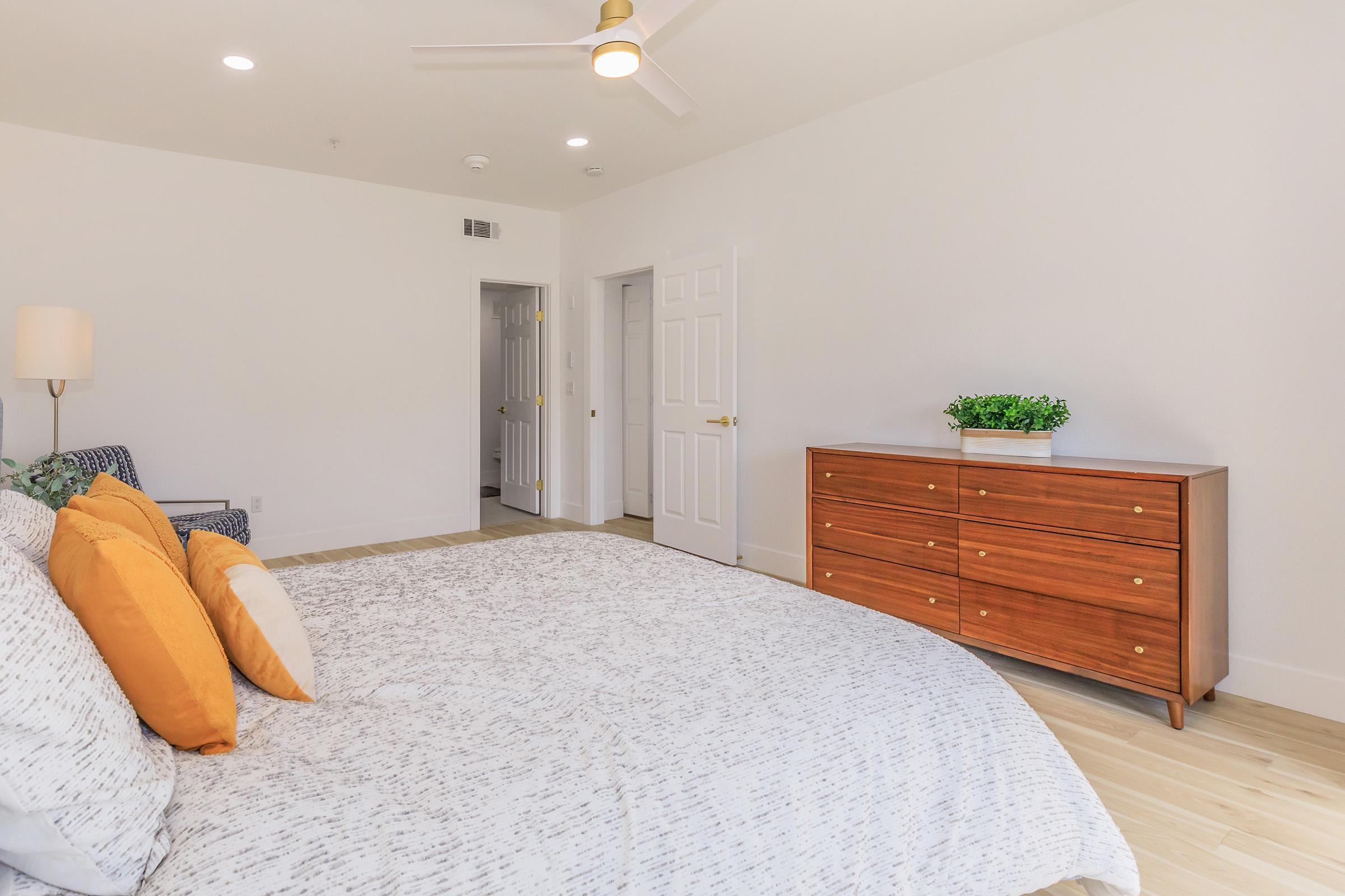 A cozy bedroom featuring a bed with gray and white bedding and orange pillows. There's a wooden dresser with a small plant on top, and a chair with a patterned fabric. The walls are light-colored, and a door leads to another room, creating a bright and inviting atmosphere.