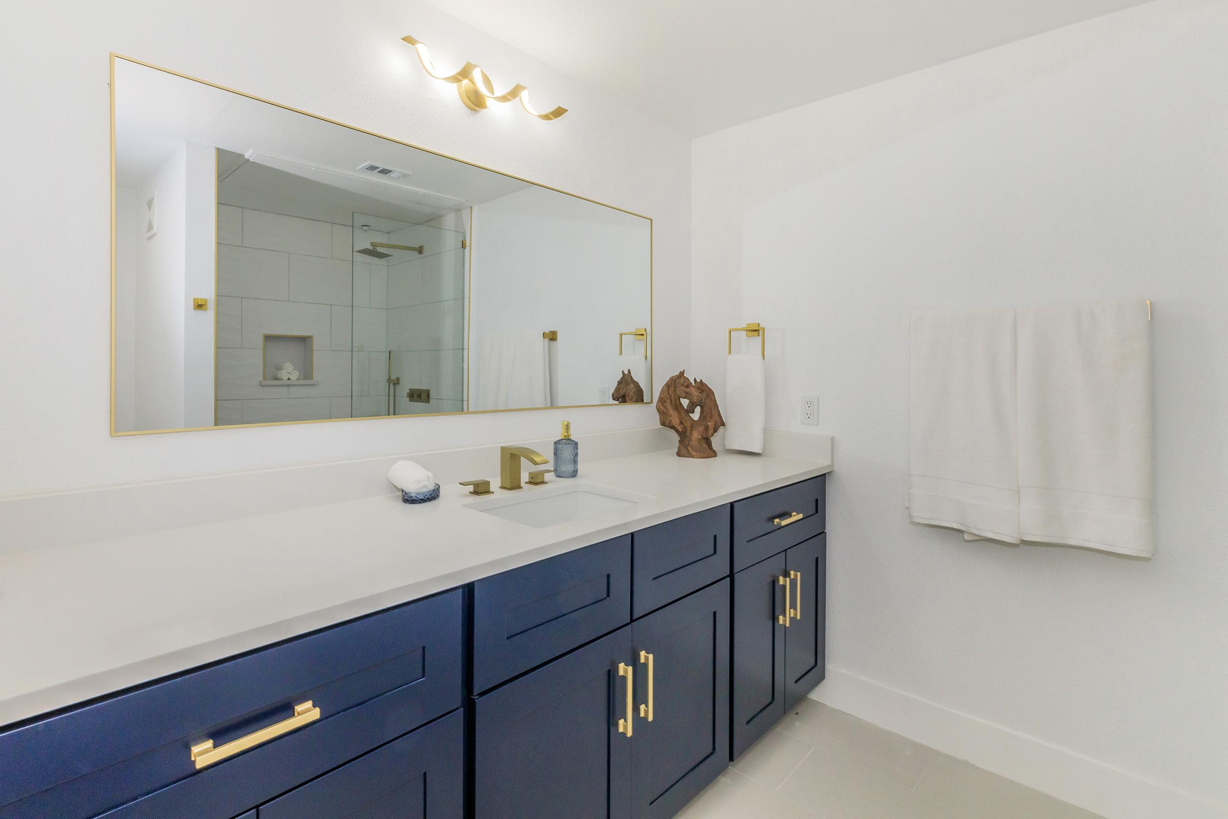 A modern bathroom featuring a large mirror with gold accents, a white countertop with a sink, and navy blue cabinetry. The space includes a glass shower stall in the background, a towel hanging on the wall, and decorative items on the countertop, creating a clean and stylish atmosphere.