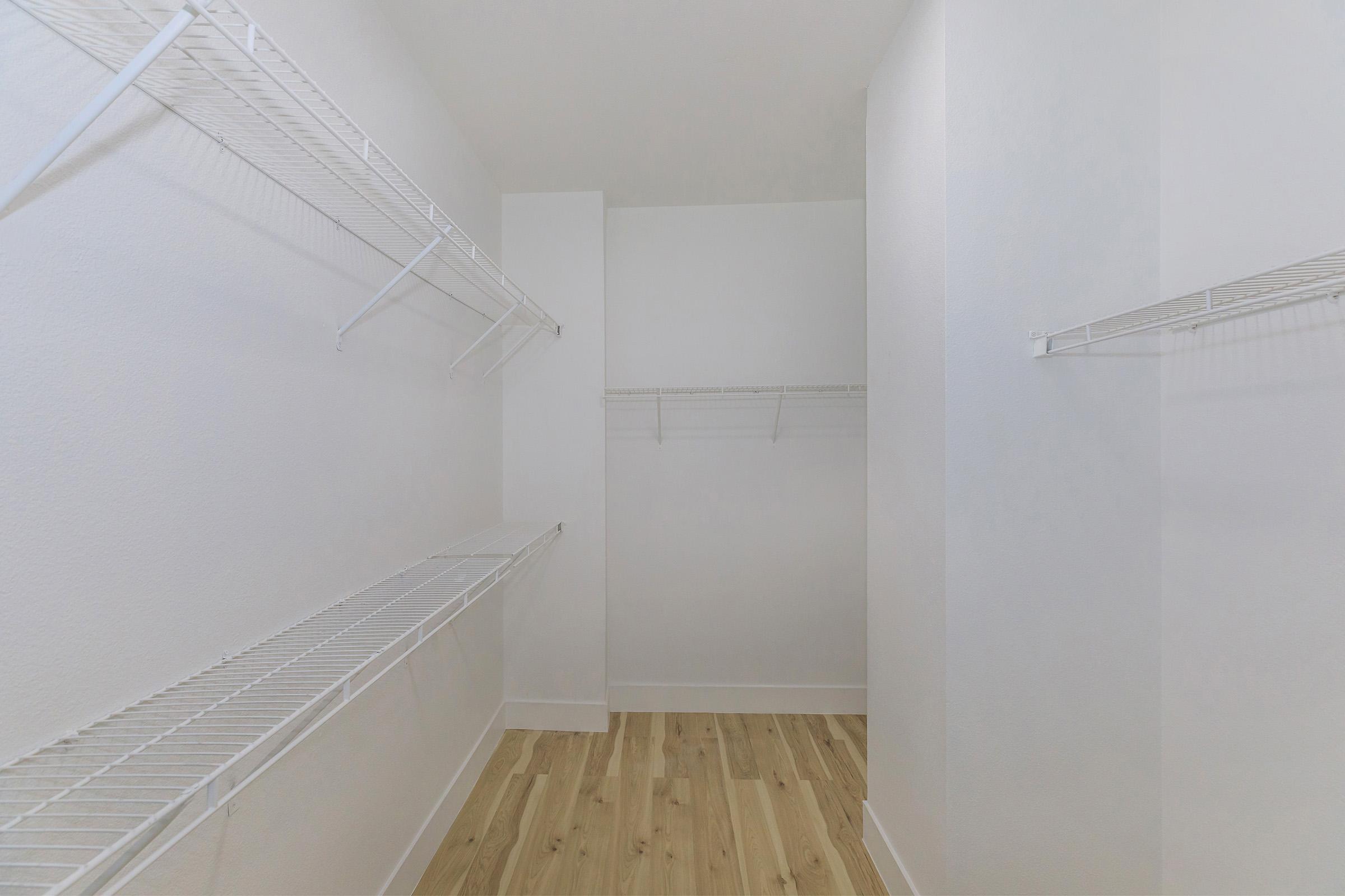 A spacious, empty closet featuring white wire shelving on two walls, with light-colored wood flooring. The walls and ceiling are painted white, creating a bright and clean appearance. The closet has ample space for organizing clothes and accessories.