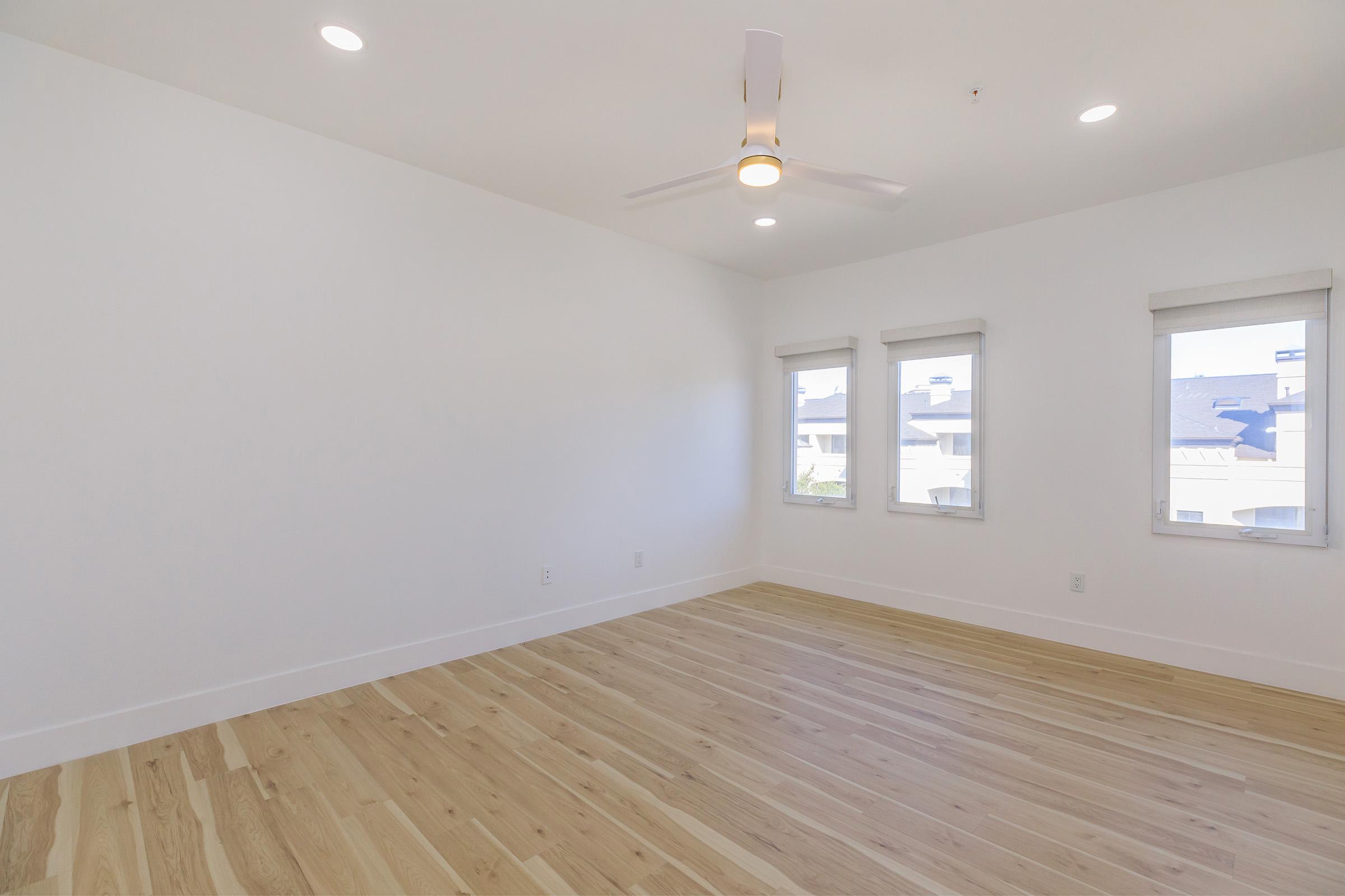 A spacious, uncluttered room with white walls and light wooden flooring. It features three windows with blinds allowing natural light, and a modern ceiling fan. The minimalist design creates a bright and airy atmosphere, ideal for diverse uses.