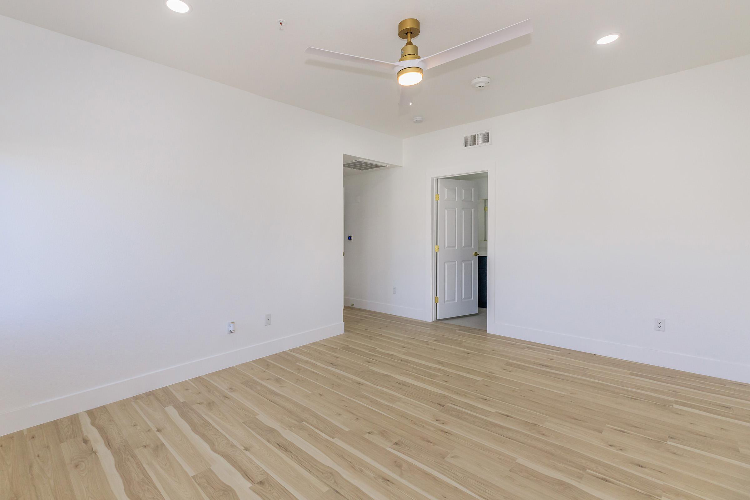 A spacious, empty room featuring light wood flooring, a ceiling fan with a gold finish, and white walls. The room has a door leading to another area, and there is soft natural light coming from the windows. The overall ambiance is bright and modern, suitable for various interior designs.
