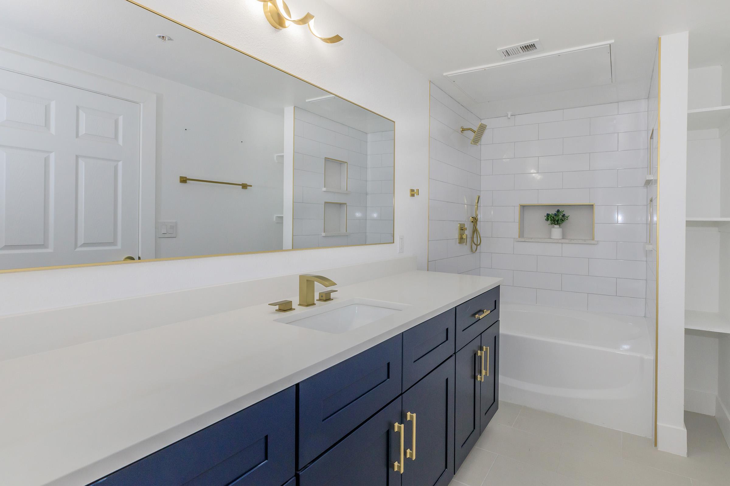 A modern bathroom featuring a navy blue vanity with gold fixtures, a white countertop, a bathtub with a showerhead, and white tiled walls. The mirror above the sink has a gold frame, and there is a small green plant displayed on the shelf. The overall design is sleek and contemporary.