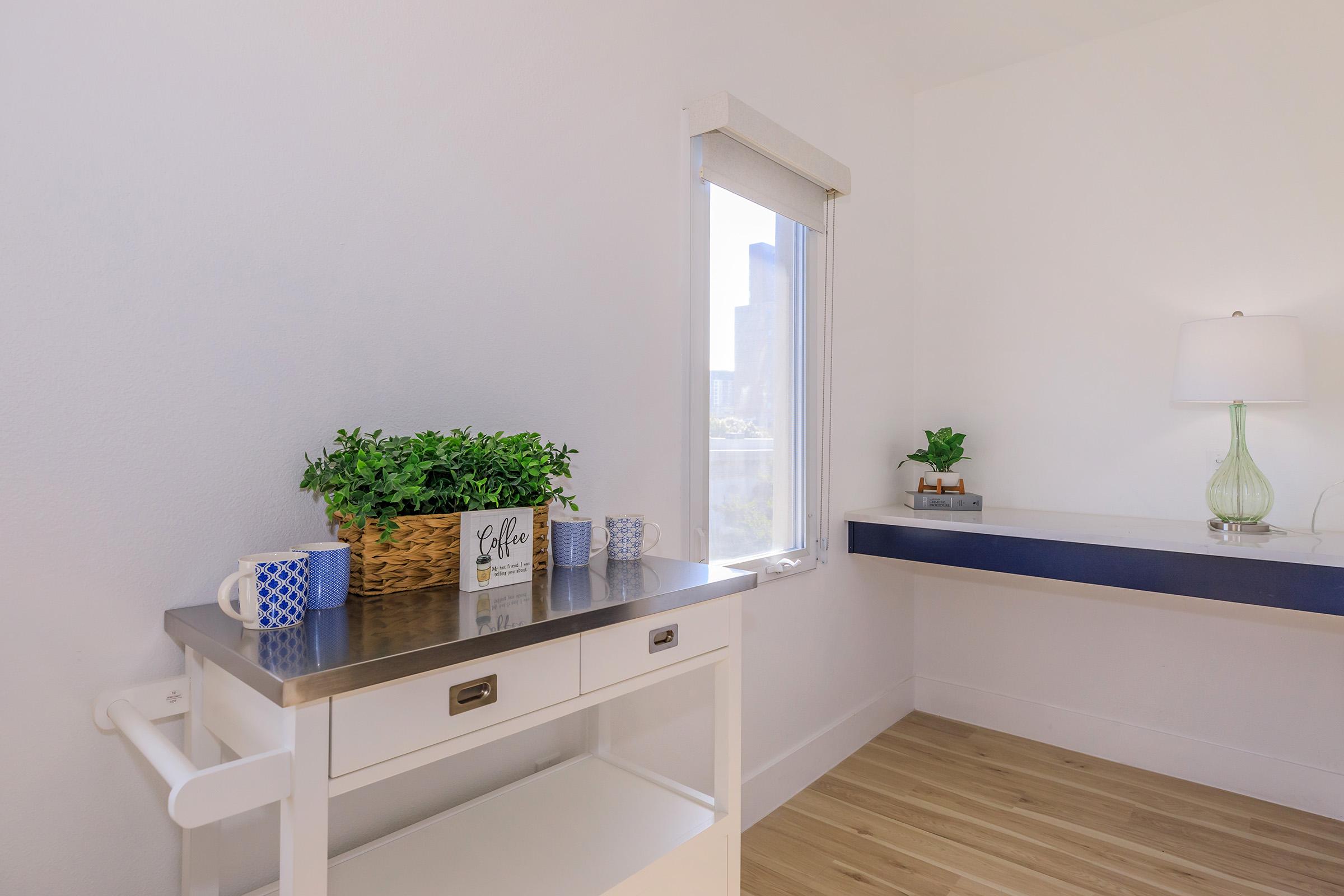 A bright, modern room featuring a white wall and wooden floor. On the left, a small white cart with a metallic top holds a blue and white mug, a decorative basket with greenery, and a sign that says "Coffee." To the right, a simple wall-mounted table with a lamp and a small plant. A window provides natural light and a city view.