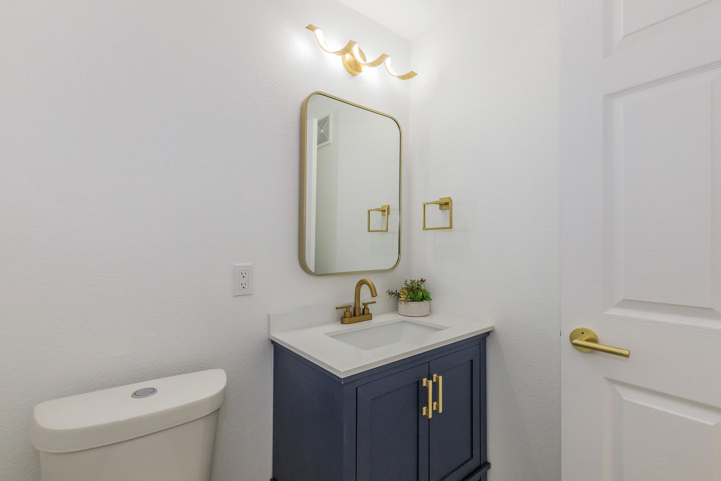 A modern bathroom featuring a white wall, a blue vanity with a sink, a round mirror mounted above, a gold faucet, a gold towel holder, and a small plant for decoration. A white toilet is visible on the left side, and the door appears to be partially open, adding light to the space.