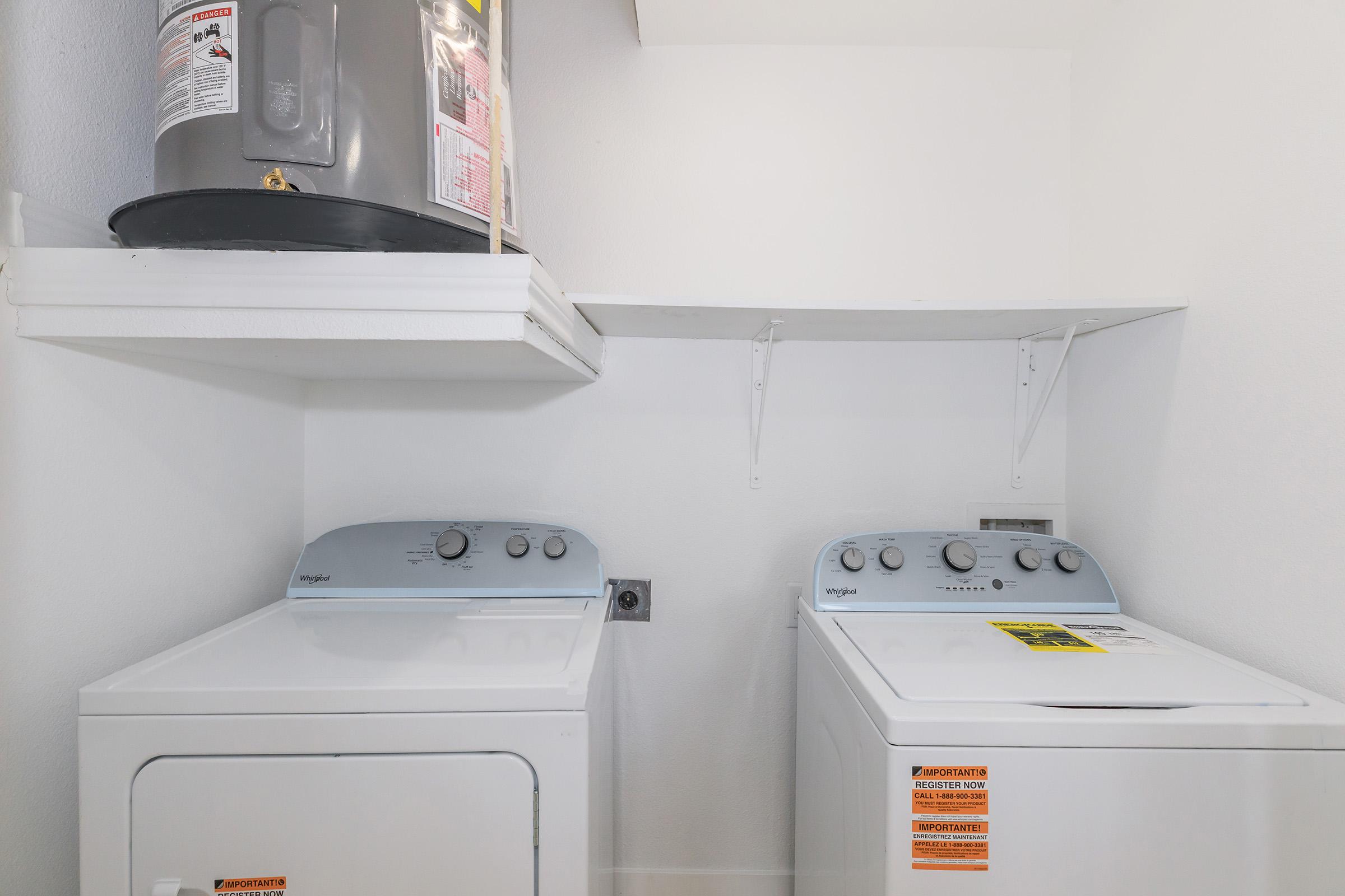 A laundry room featuring a washing machine and a dryer side by side, both labeled with safety warnings. Above them, there is a shelf and a water heater mounted on the wall. The walls are painted white, providing a bright and clean appearance.