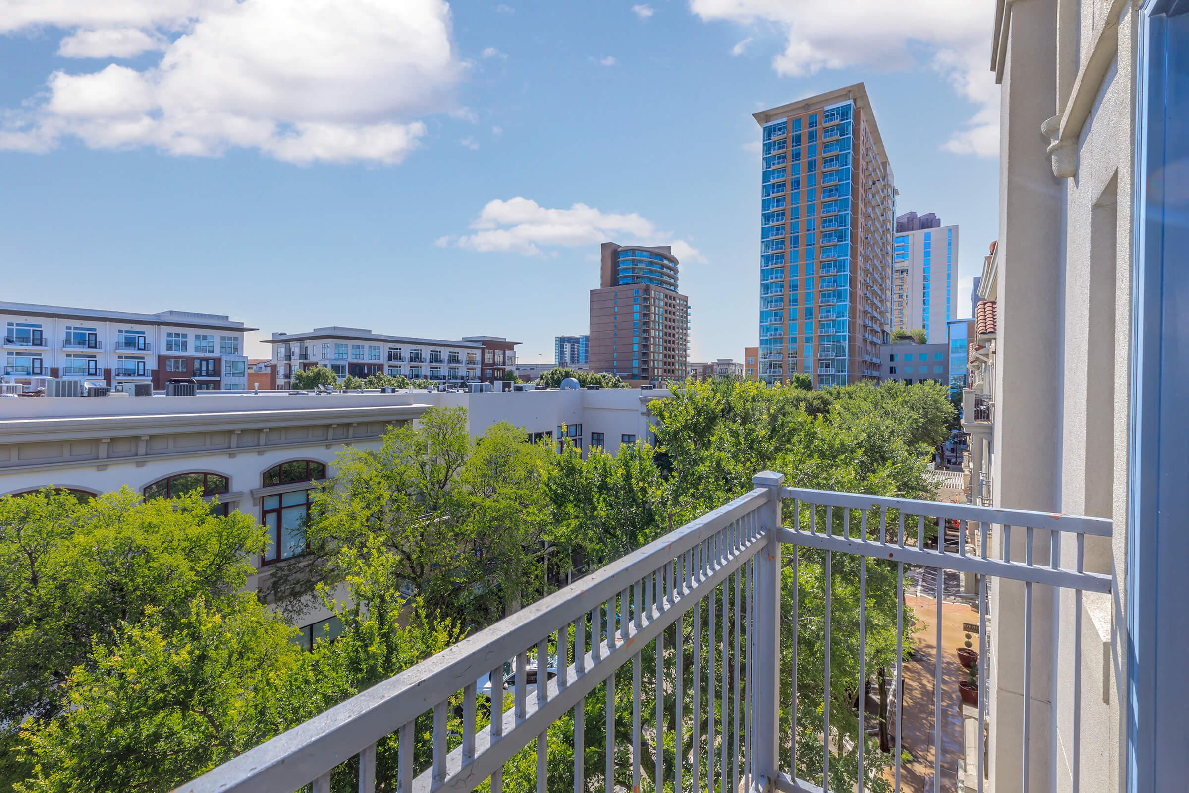 View from a balcony overlooking a vibrant cityscape. Trees line the street below, with modern buildings and high-rises in the distance under a clear blue sky with fluffy clouds. The scene captures a blend of urban architecture and greenery, showcasing a lively neighborhood atmosphere.