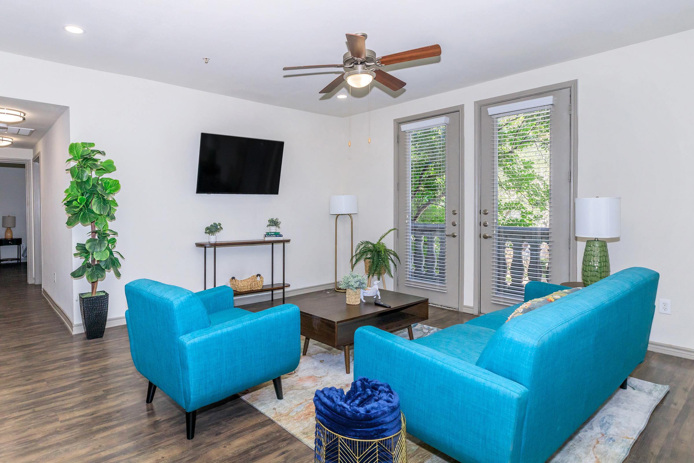 Modern living room featuring a light blue couch and armchair, a wooden coffee table, and decorative plants. A flat-screen TV is mounted on the wall, and large windows with blinds allow natural light. The flooring is dark wood, enhancing the contemporary design of the space.