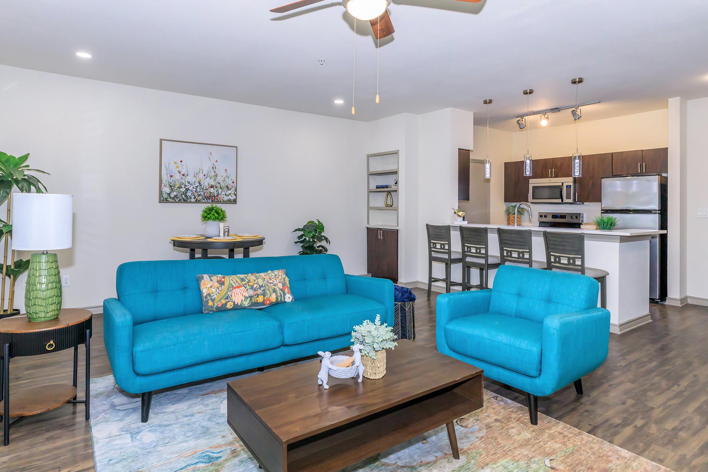 A modern living room featuring a turquoise sofa and armchair, a wooden coffee table, and decorative plants. In the background, a kitchen with dark cabinetry and stainless steel appliances is visible. The space is well-lit, creating a welcoming atmosphere.