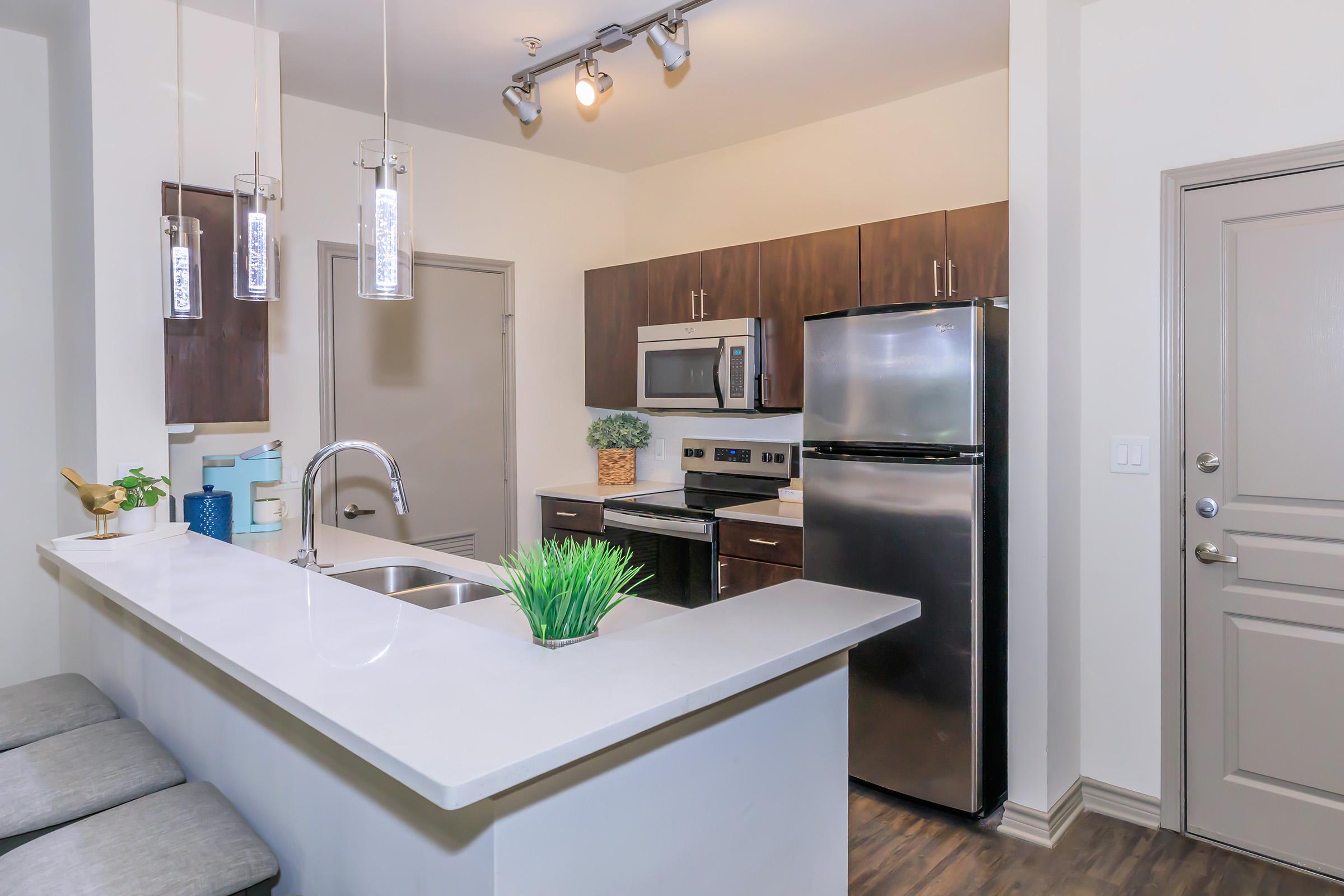 A modern kitchen featuring a gray countertop with a sink, stainless steel appliances including a refrigerator and microwave, dark wood cabinets, and pendant lighting. A small potted plant adds a touch of greenery, and there are stools at the counter. The space is well-lit and designed for a contemporary look.