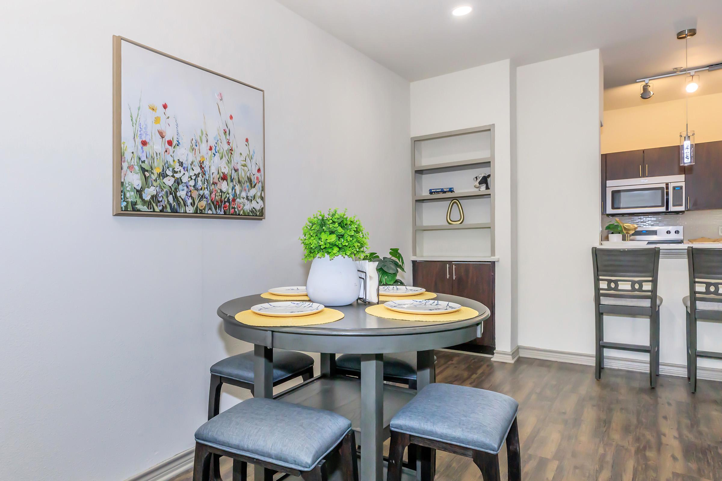 A dining area featuring a round table with four chairs, adorned with yellow placemats and decorative plates. A vibrant floral painting hangs on the wall, while an open kitchen with modern appliances is visible in the background. A potted plant adds a touch of greenery to the scene.