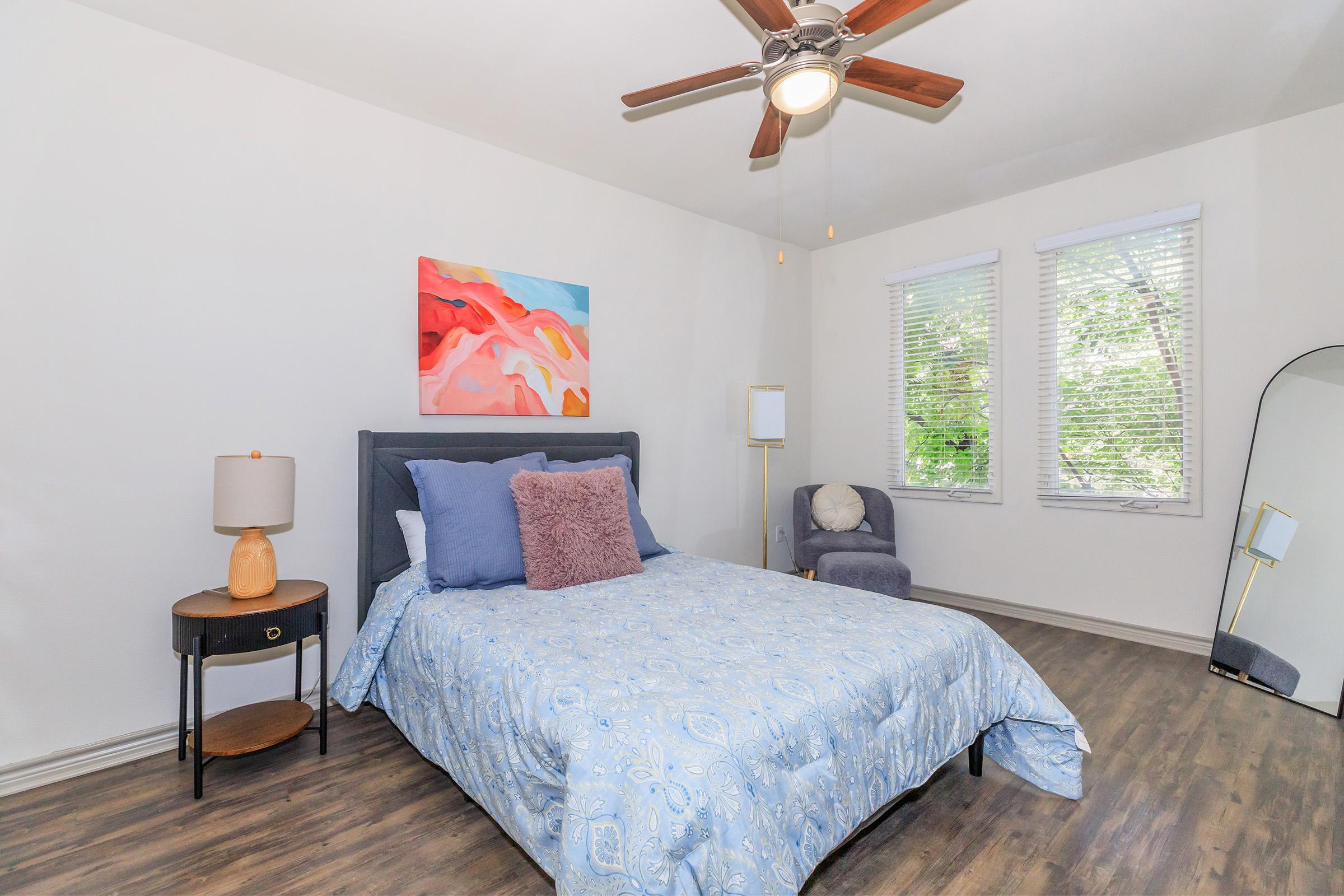 A cozy bedroom featuring a queen-sized bed with a light blue patterned blanket and decorative pillows. A bedside table with a lamp sits next to the bed, while a plush gray chair and a full-length mirror are present. The room has white walls, large windows with blinds, and a colorful abstract wall painting.