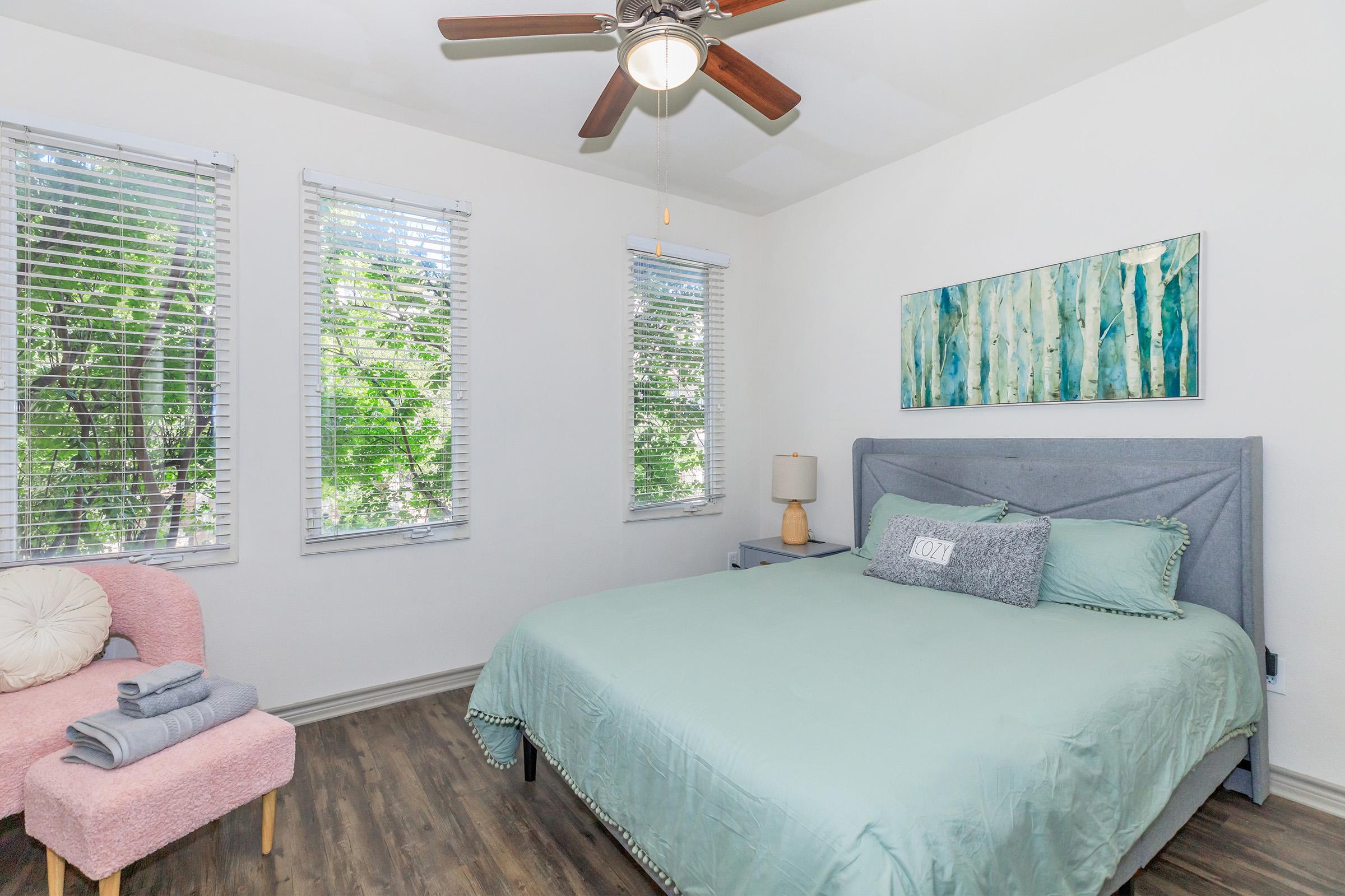 A bright, modern bedroom featuring a king-size bed with a light teal comforter and decorative pillows. A small pink chair is in the corner, and there's a lamp on a nightstand beside the bed. Large windows with blinds allow natural light to fill the room, and a piece of abstract wall art adds a touch of color.