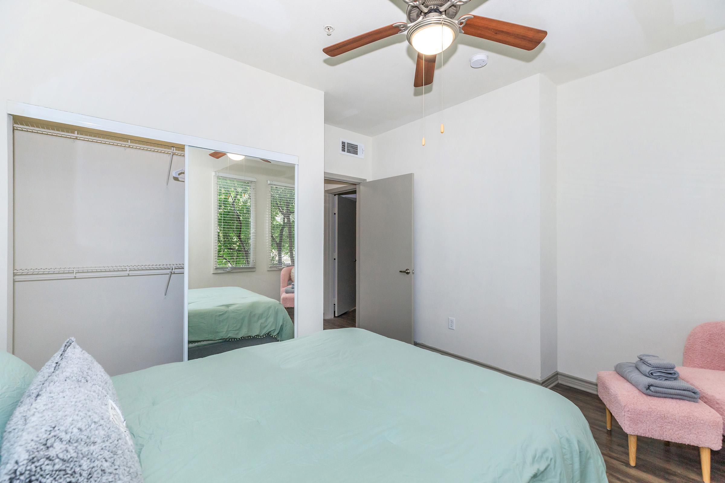 A neatly arranged bedroom featuring a green bedspread, a ceiling fan, and natural light coming through a window. There is a closet with mirrored doors, a door leading to another room, and a pink chair next to a neatly stacked set of towels. The walls are painted in a light color.