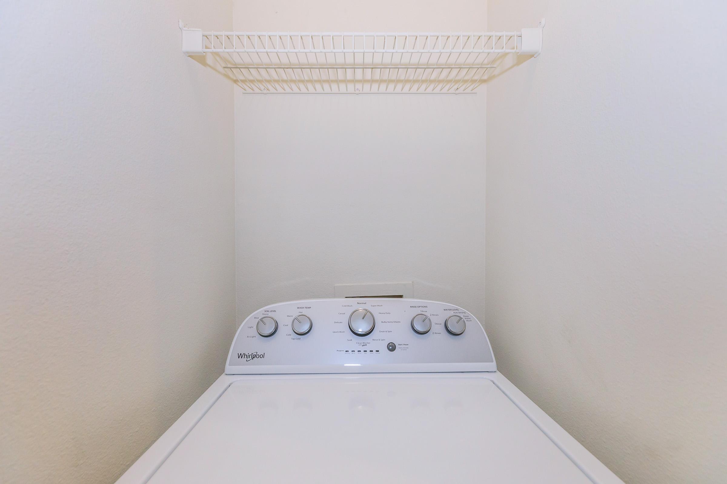 A white washing machine with a control panel featuring several dials and buttons, positioned in a small laundry space. Above the machine, there is a wire shelf against a plain wall, providing storage space. The scene is well-lit and organized, suggesting a tidy laundry area.