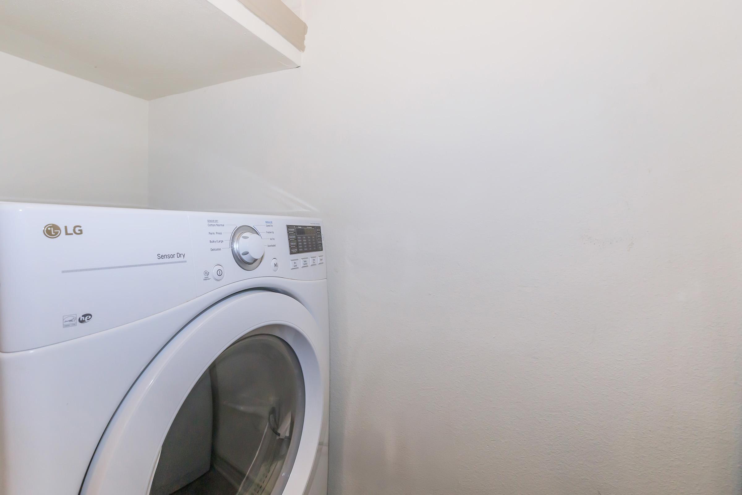 A white LG dryer is positioned in a small laundry space. The dryer features a control panel with various settings and is placed against a plain, light-colored wall. A shelf is visible above the dryer, with no items placed on it. The overall atmosphere is clean and functional.