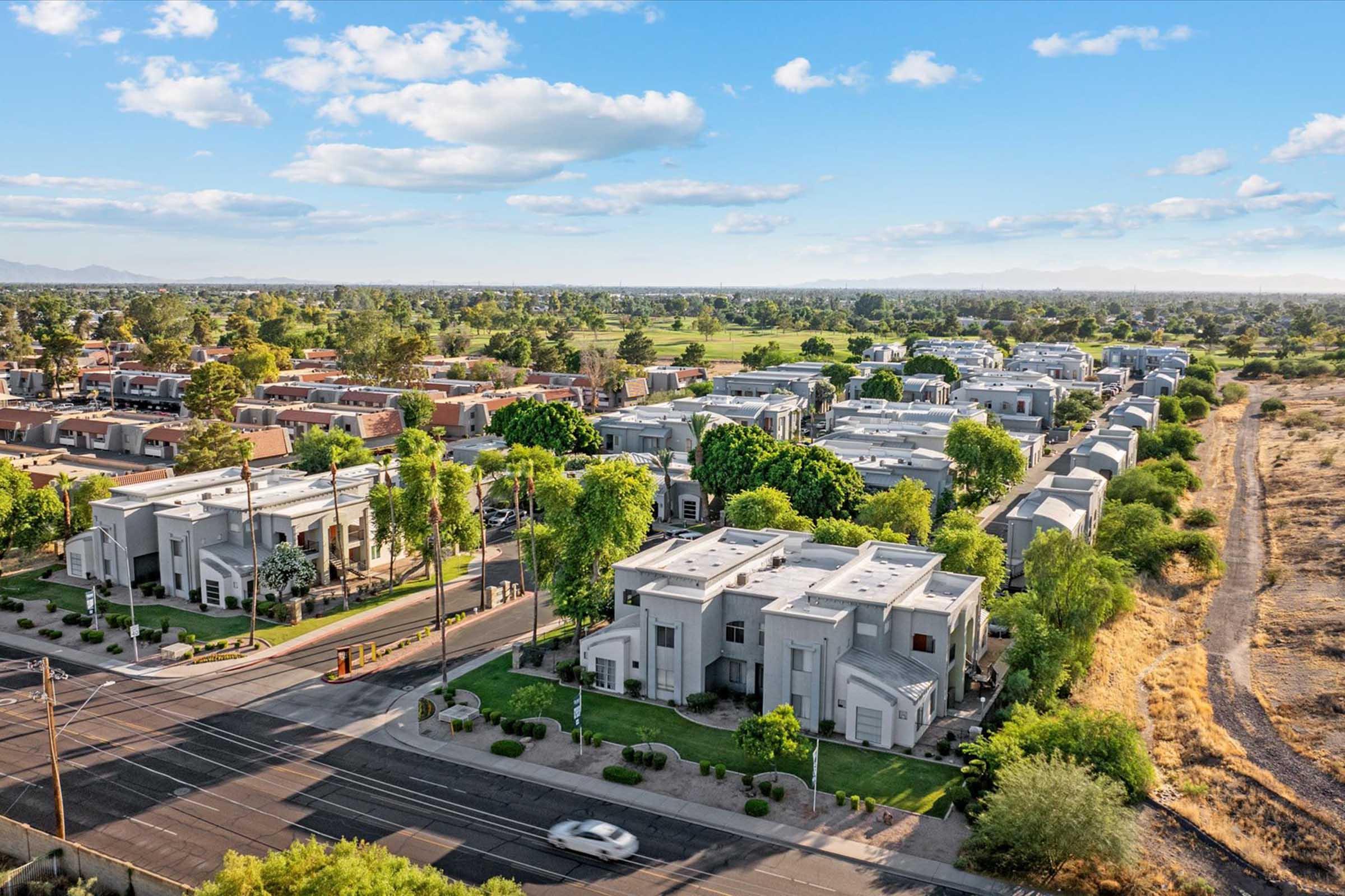 Aerial view of a residential area featuring modern houses surrounded by greenery and trees. A road runs along the front, with cars visible. In the background, open fields and additional homes extend towards the horizon under a partly cloudy sky.