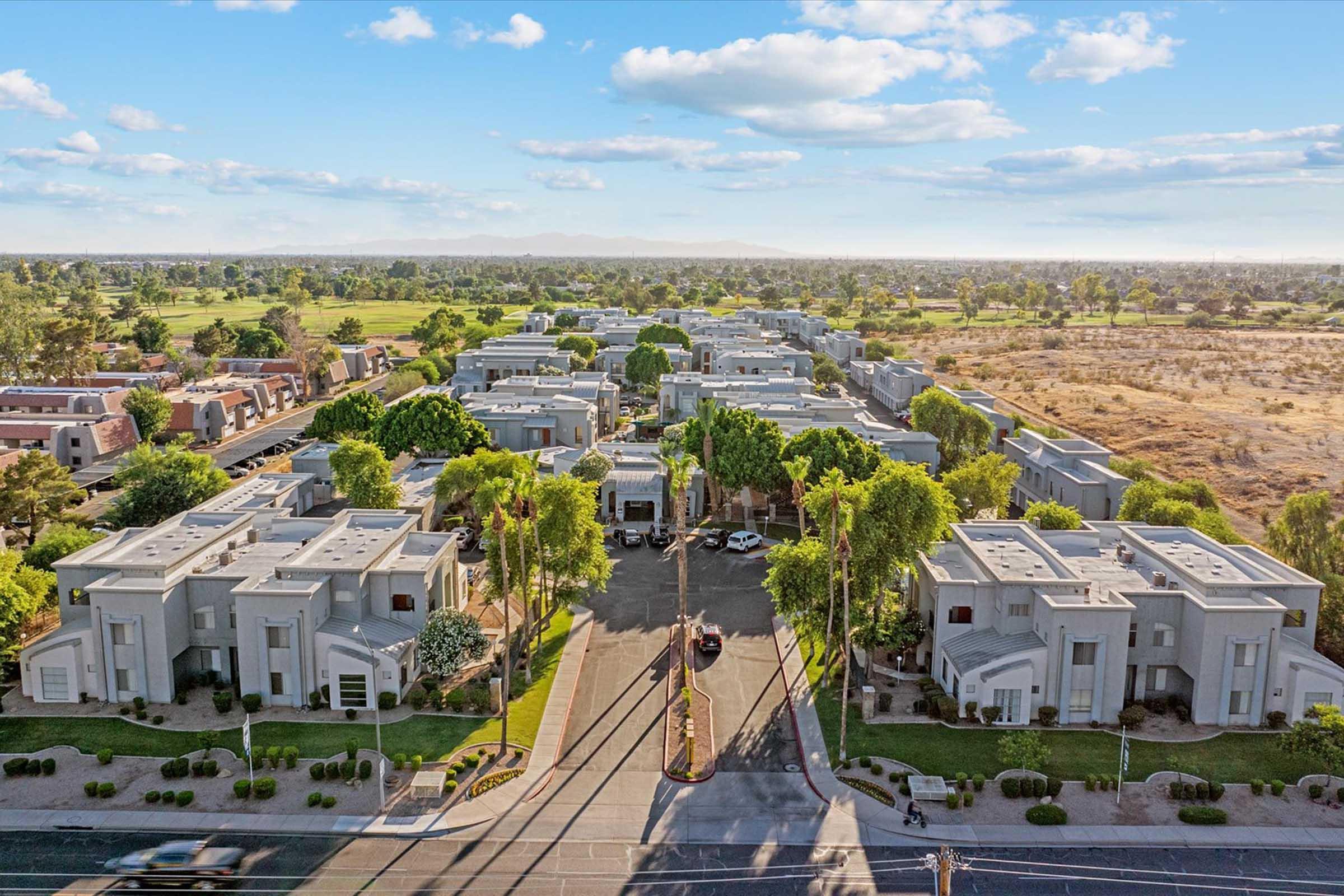Aerial view of a residential area featuring modern, low-rise buildings surrounded by greenery. The scene includes a wide road with parked cars, leading to landscaped lawns and palm trees, set against a backdrop of open fields and distant mountains under a clear blue sky.