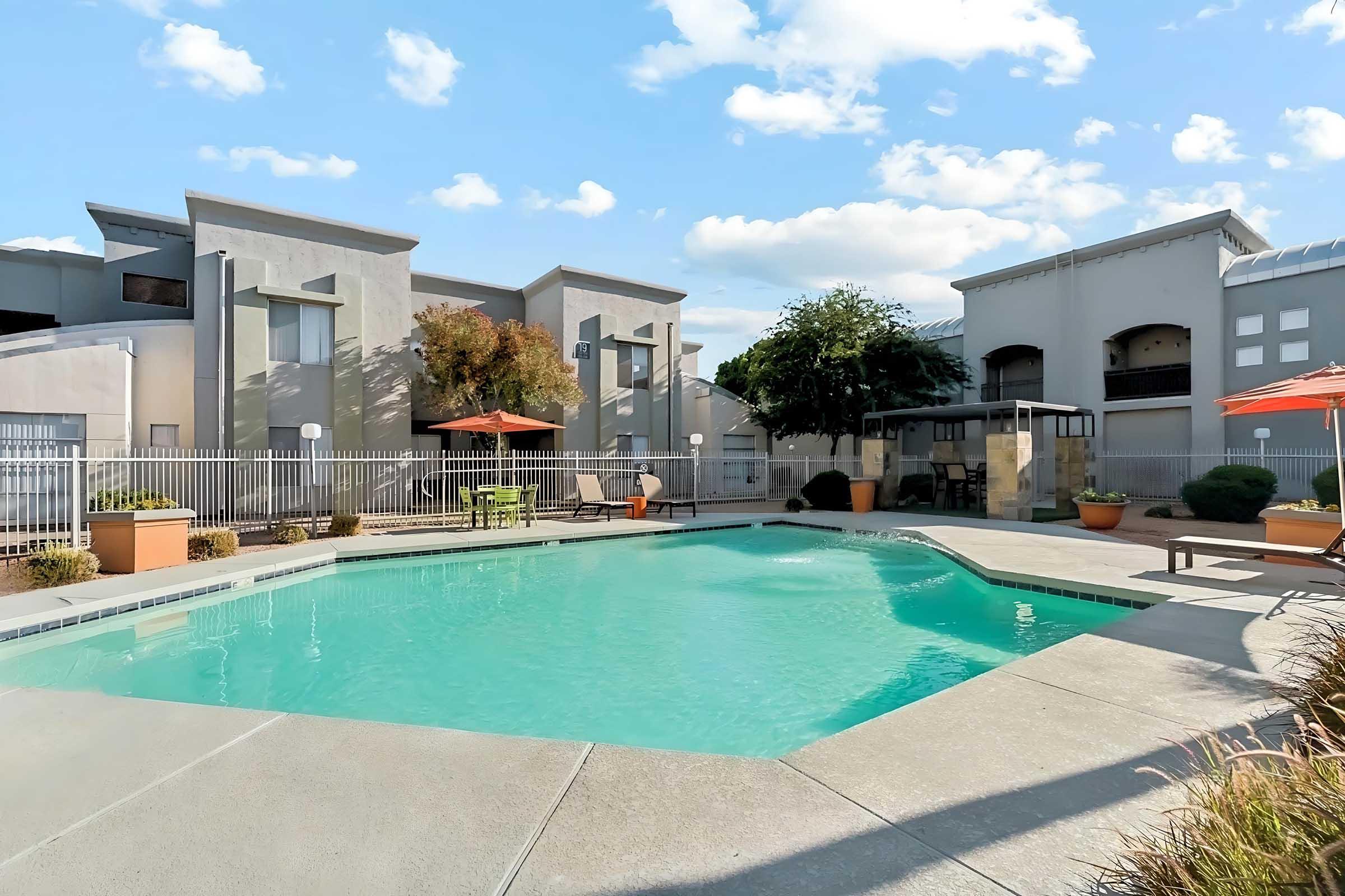 A clear swimming pool surrounded by a concrete deck, with lounge chairs and orange umbrellas. Modern apartment buildings are in the background, and the sky is bright with scattered clouds. Potted plants add greenery to the space, creating a relaxing outdoor environment.