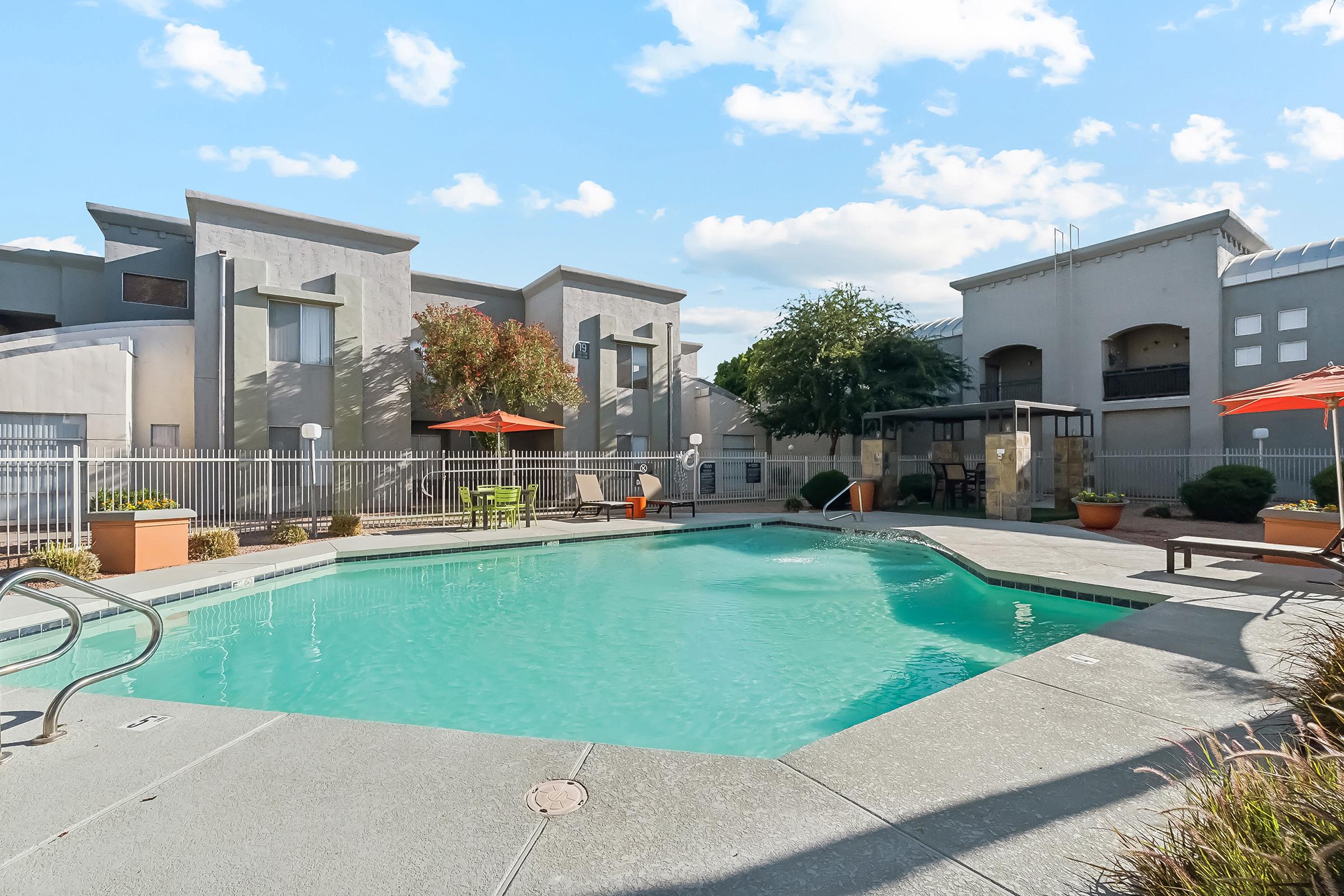A serene swimming pool area featuring a clear blue pool surrounded by concrete decking. There are lounge areas with orange umbrellas and potted plants. Modern apartment buildings form the backdrop under a bright sky with scattered clouds.