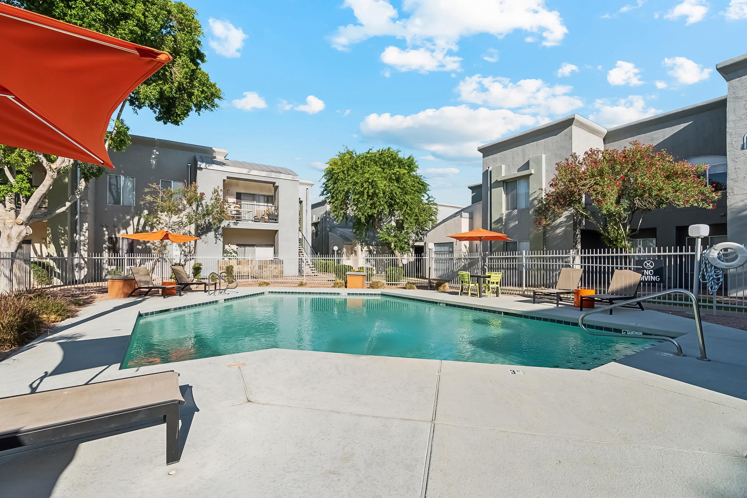 A serene pool area surrounded by apartment buildings, featuring lounge chairs and orange umbrellas. Lush green trees provide shade, and a fenced perimeter adds privacy. The clear blue sky with scattered clouds complements the inviting atmosphere.