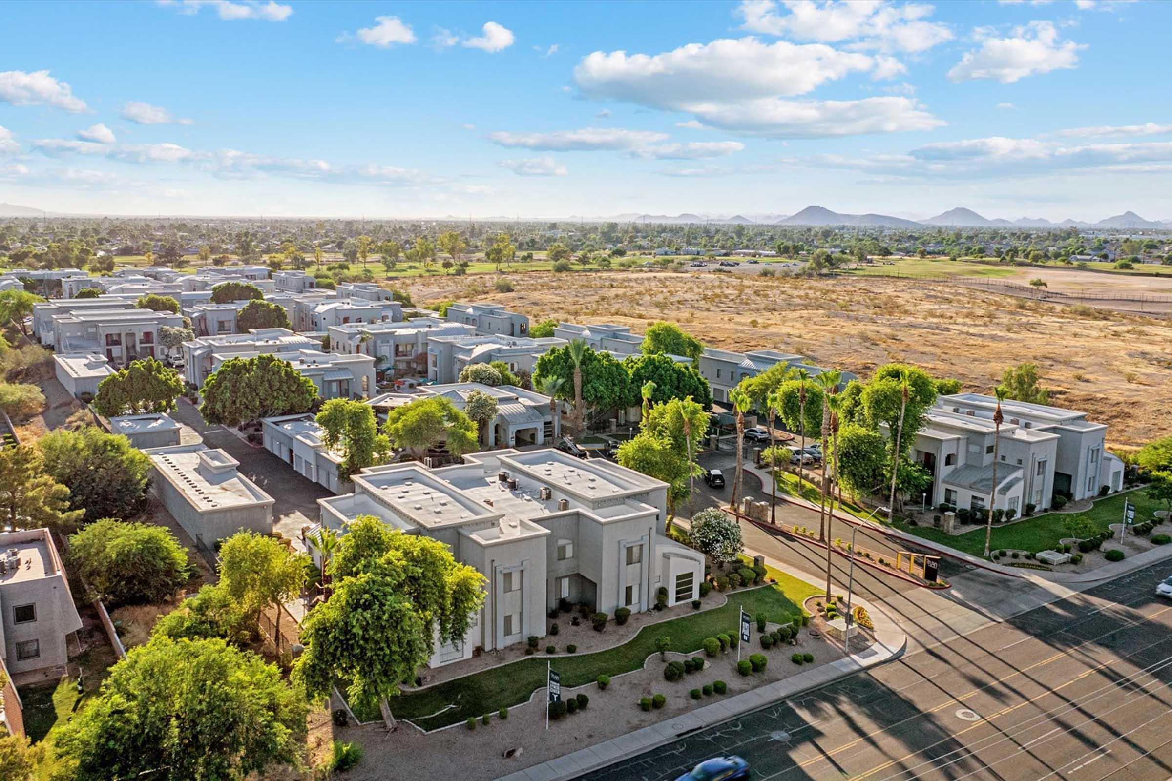 Aerial view of a suburban neighborhood featuring modern, single-story homes surrounded by greenery. The area is dotted with trees, and a road runs along the front. In the background, open land and distant mountains are visible under a blue sky with fluffy clouds.