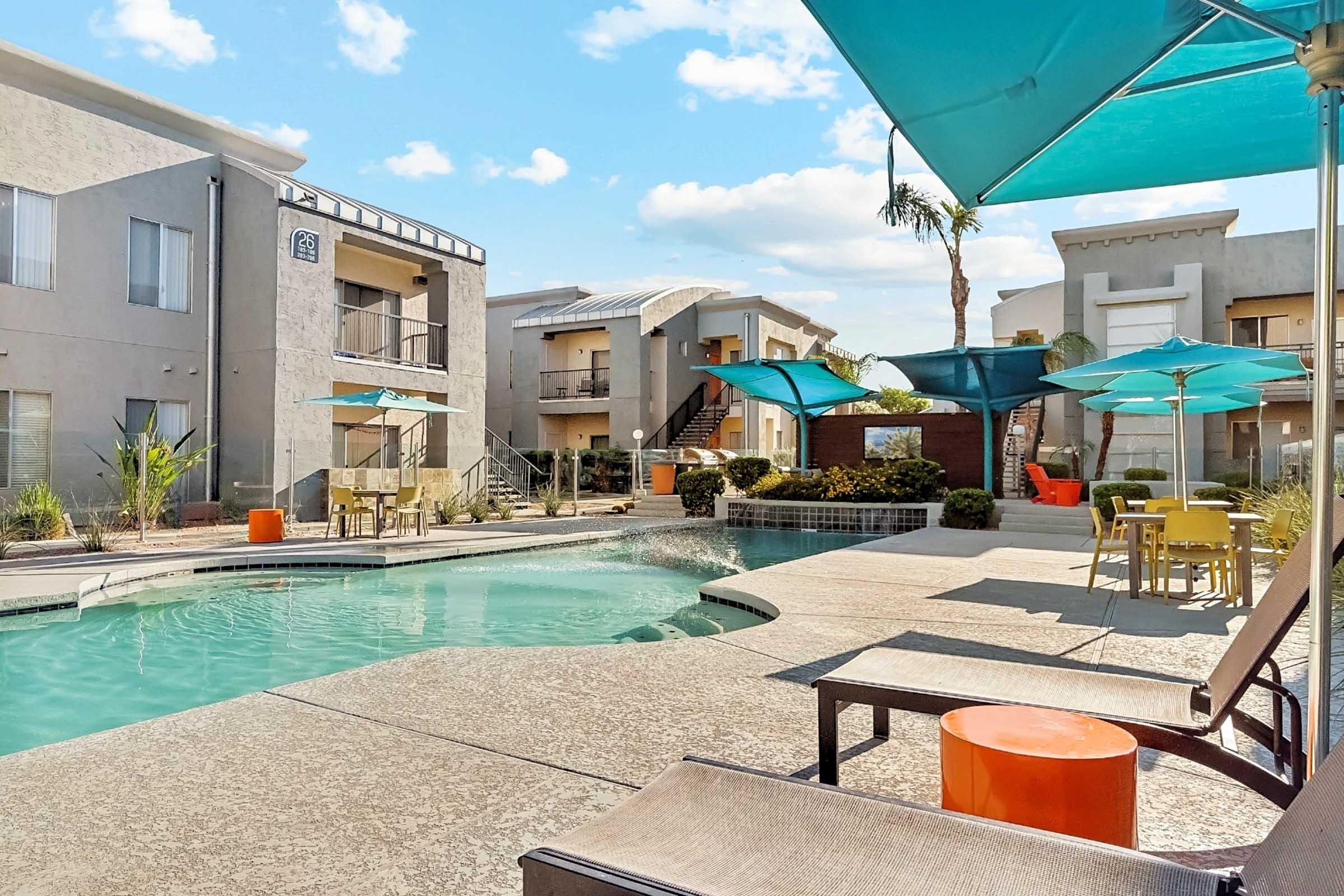 A relaxing pool area at an apartment complex, featuring lounge chairs and colorful umbrellas. The pool is surrounded by landscaped greenery, and modern buildings are visible in the background under a bright blue sky with fluffy clouds. A vibrant outdoor seating area is also present.
