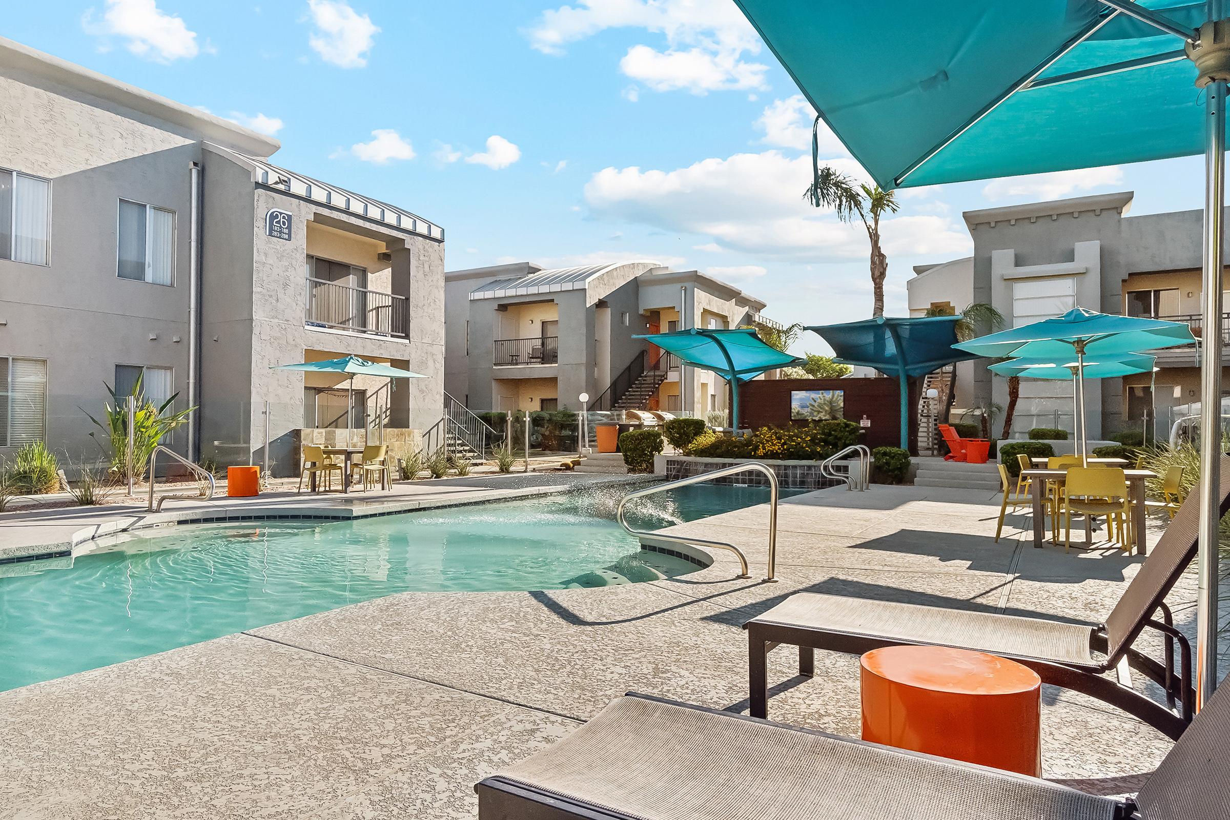 A tranquil pool area surrounded by modern apartment buildings. Lounge chairs and colorful umbrellas provide shade, while palm trees add a touch of greenery. Tables and chairs set nearby create a relaxed atmosphere for residents to enjoy the outdoor space.