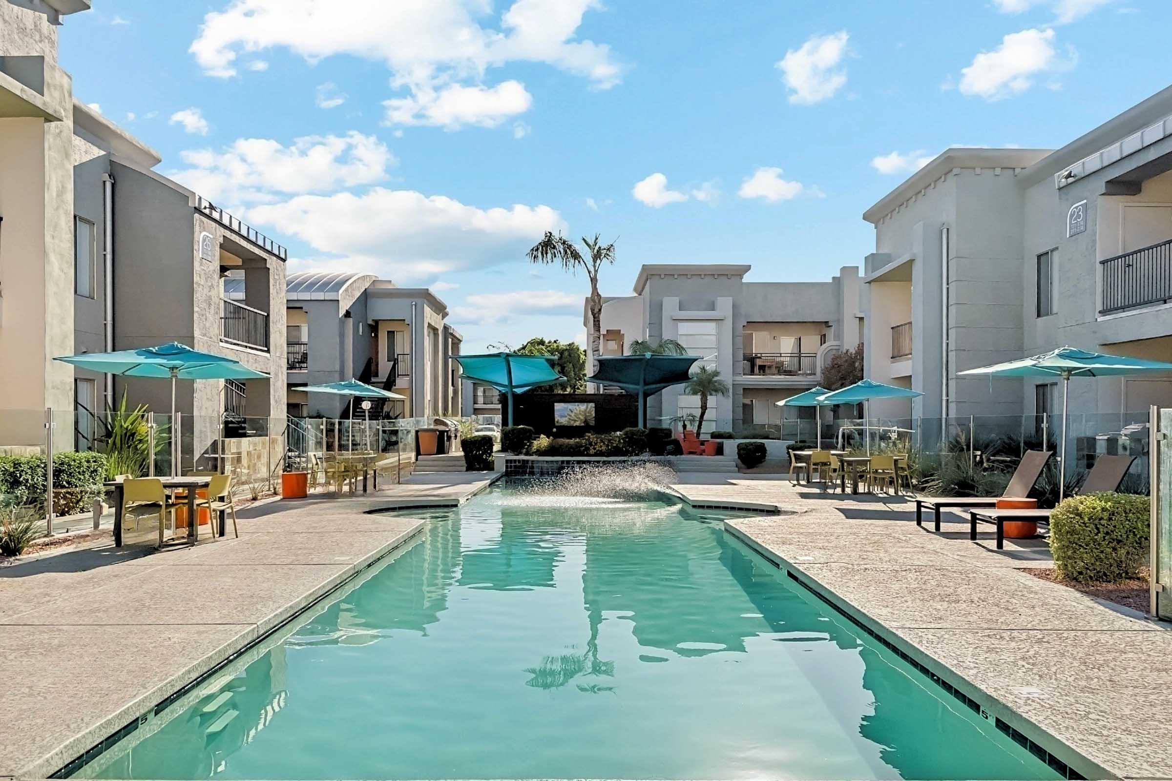 A serene apartment complex featuring a swimming pool surrounded by lounge chairs and tables with umbrellas. Lush greenery and palm trees are visible, creating a relaxing atmosphere. The sky is clear with a few fluffy clouds, enhancing the inviting outdoor space.
