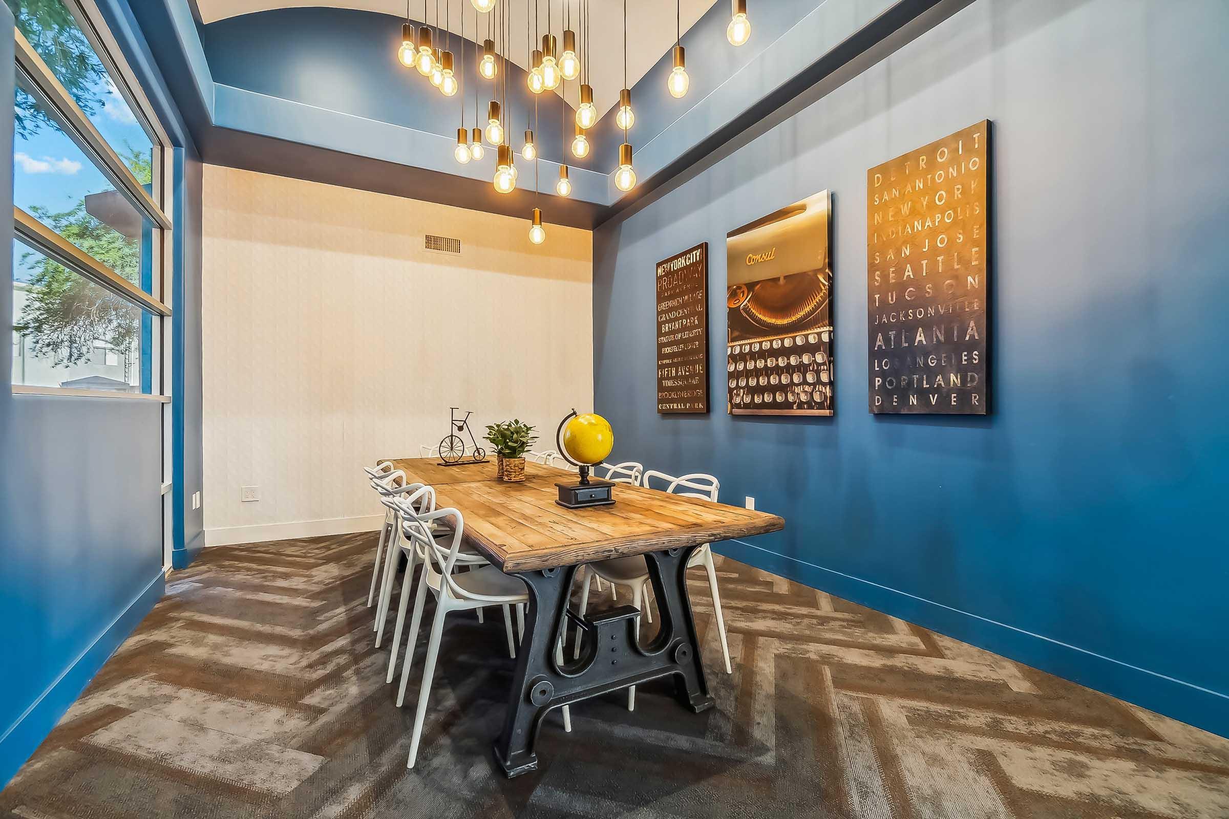 A modern conference room featuring a rustic wooden table surrounded by white chairs. The walls are painted a vibrant blue and adorned with framed art. Pendant lights hang from the ceiling, creating a warm atmosphere. A small potted plant and a globe are on the table, adding a touch of decor.