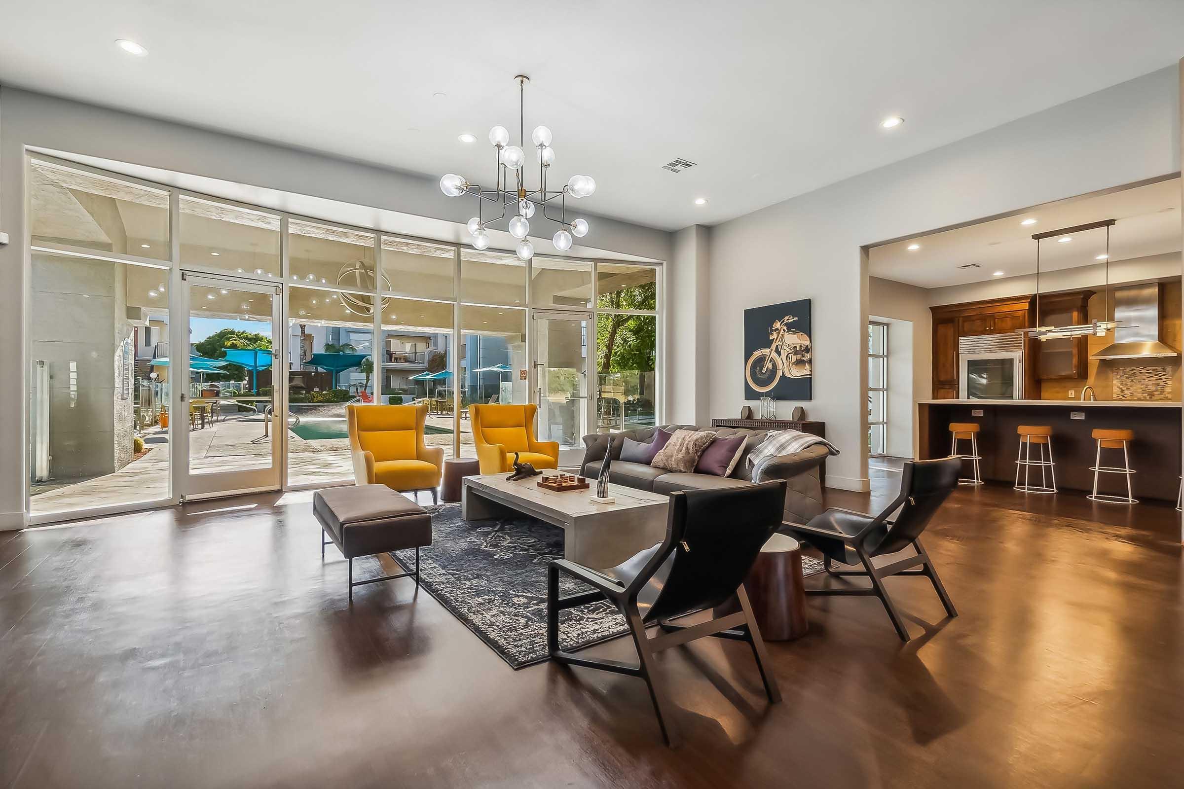 A modern living room featuring large glass windows, two yellow armchairs, a dark sectional sofa, and an elegant coffee table. There’s a patterned area rug and a stylish chandelier above. In the background, there’s a view of an outdoor space with seating and an accessible kitchen bar area.