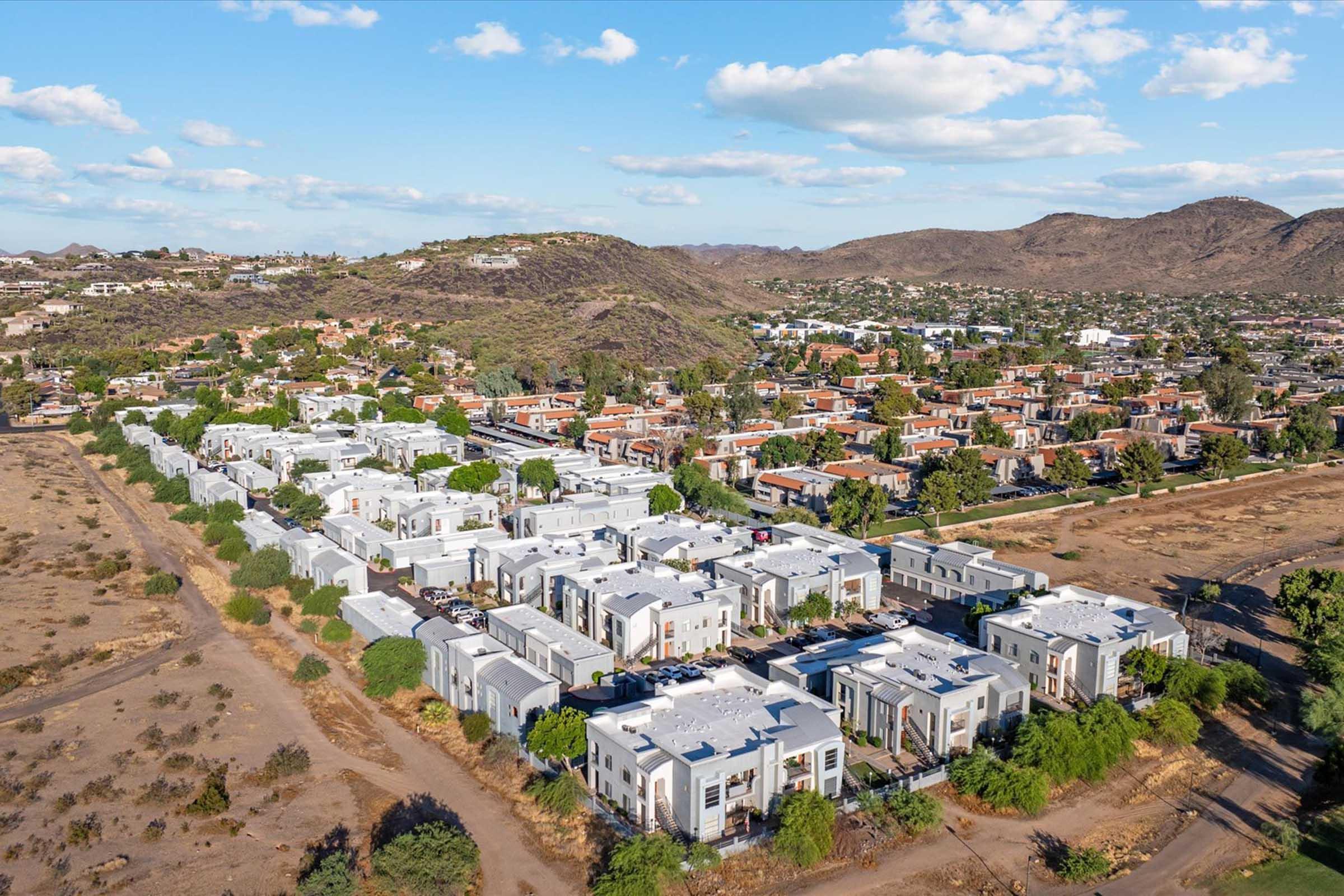 Aerial view of a residential area featuring modern buildings with flat roofs, surrounded by sparse vegetation and hills in the background. The landscape transitions from developed neighborhoods to open, natural terrain under a partly cloudy sky.